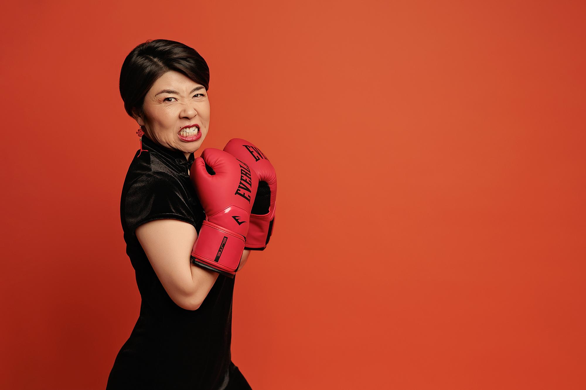 Powerful personal branding photography Calgary: Fierce woman in a black velvet qipao and red tassel earrings holding red boxing gloves in a guard stance with an intense expression against a bold red background