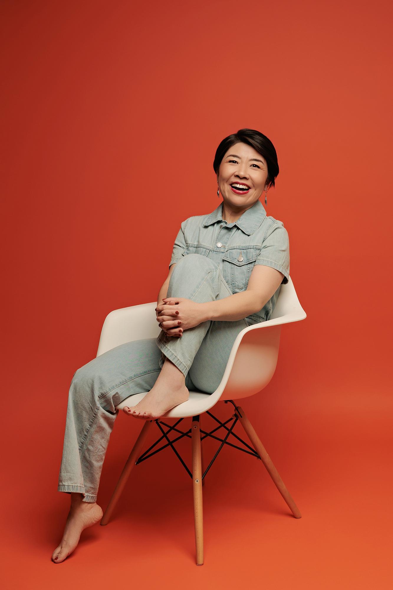 Joyful personal branding photography Calgary studio: Radiant woman with short dark hair in a light denim short-sleeve jumpsuit laughing with one knee pulled up while seated in a white Eames chair, bold red-orange background