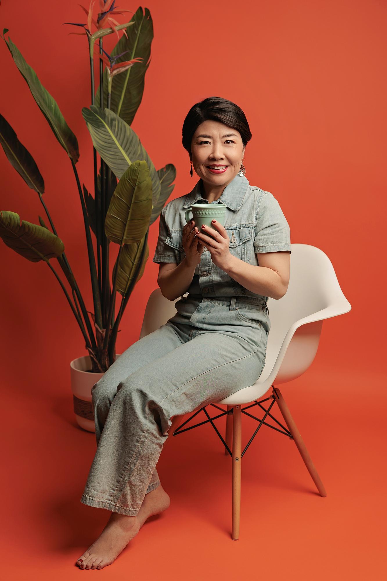 Lifestyle personal branding Calgary studio: Relaxed woman with short dark hair in a light denim jumpsuit seated in a white Eames chair holding a sage green mug, a tropical bird-of-paradise plant beside her against a red-orange background