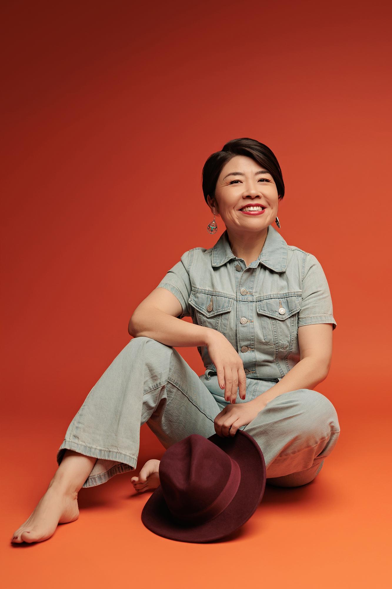 Fun personal branding photography Calgary studio: Joyful woman with short dark hair in a light denim jumpsuit seated barefoot on the studio floor laughing upward while holding a burgundy hat, red-orange background