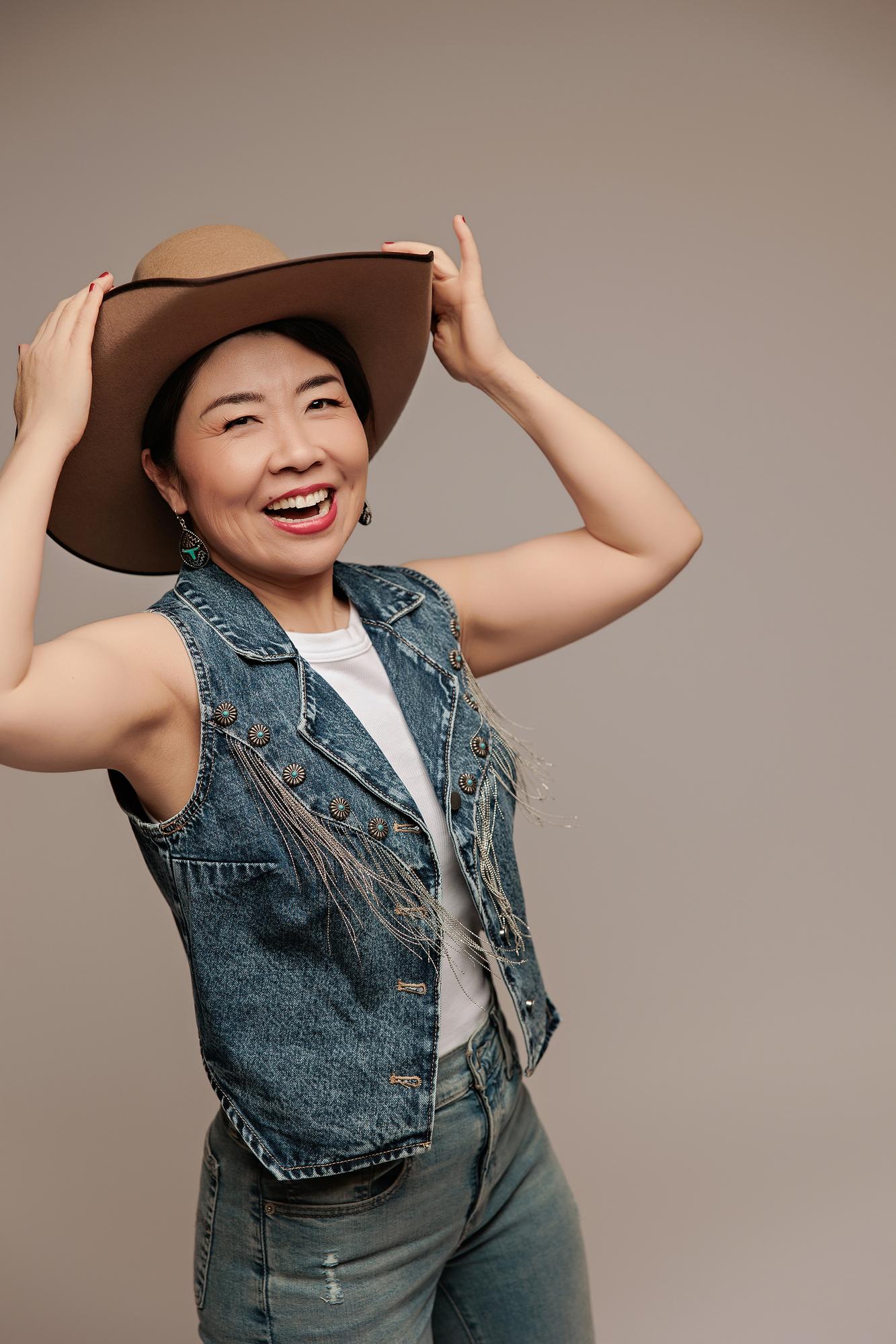 Vibrant personal branding headshot Calgary studio: Energetic woman with short dark hair laughing while holding a tan wide-brim cowboy hat overhead with both hands, wearing a studded denim vest and jeans against a warm taupe background