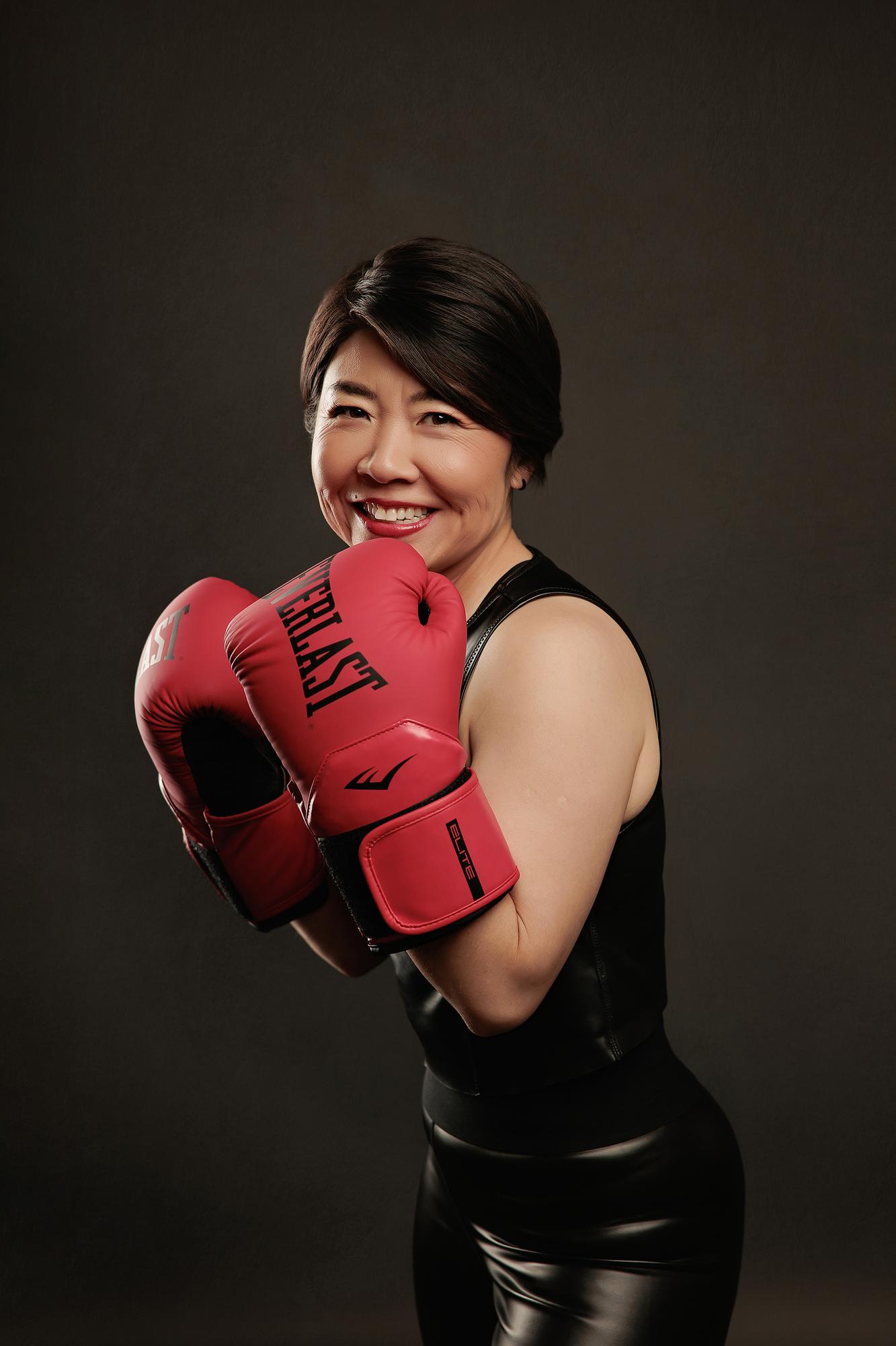 Dynamic personal branding photography Calgary: Joyful woman in a black sleeveless top and leather pants posing with red Everlast boxing gloves in a fighting stance, dark studio background