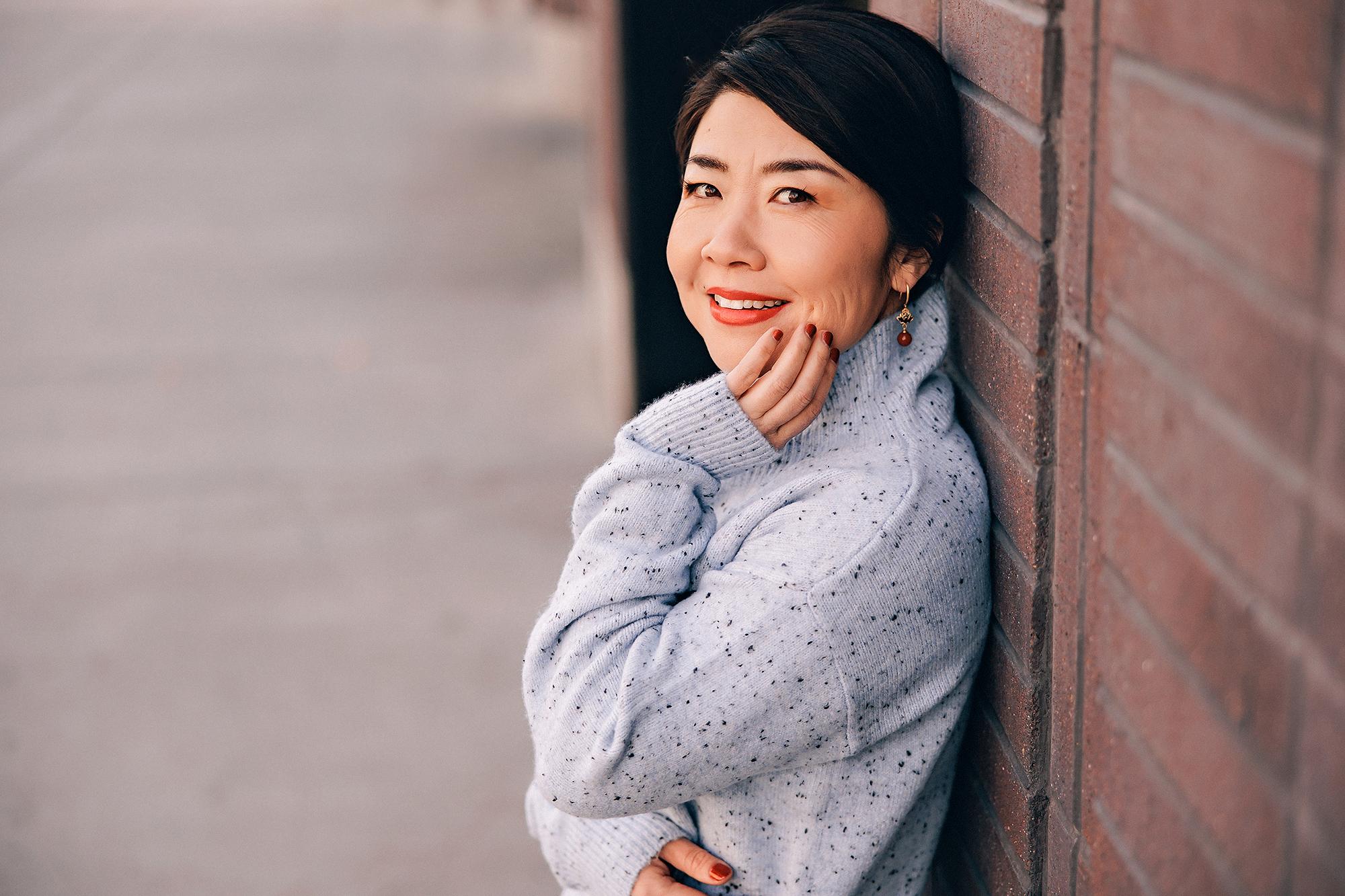 On-location personal branding headshot Calgary: Warm close-up of a woman with short dark hair in a grey speckled turtleneck sweater leaning against an exposed brick wall with hand on cheek and a soft smile