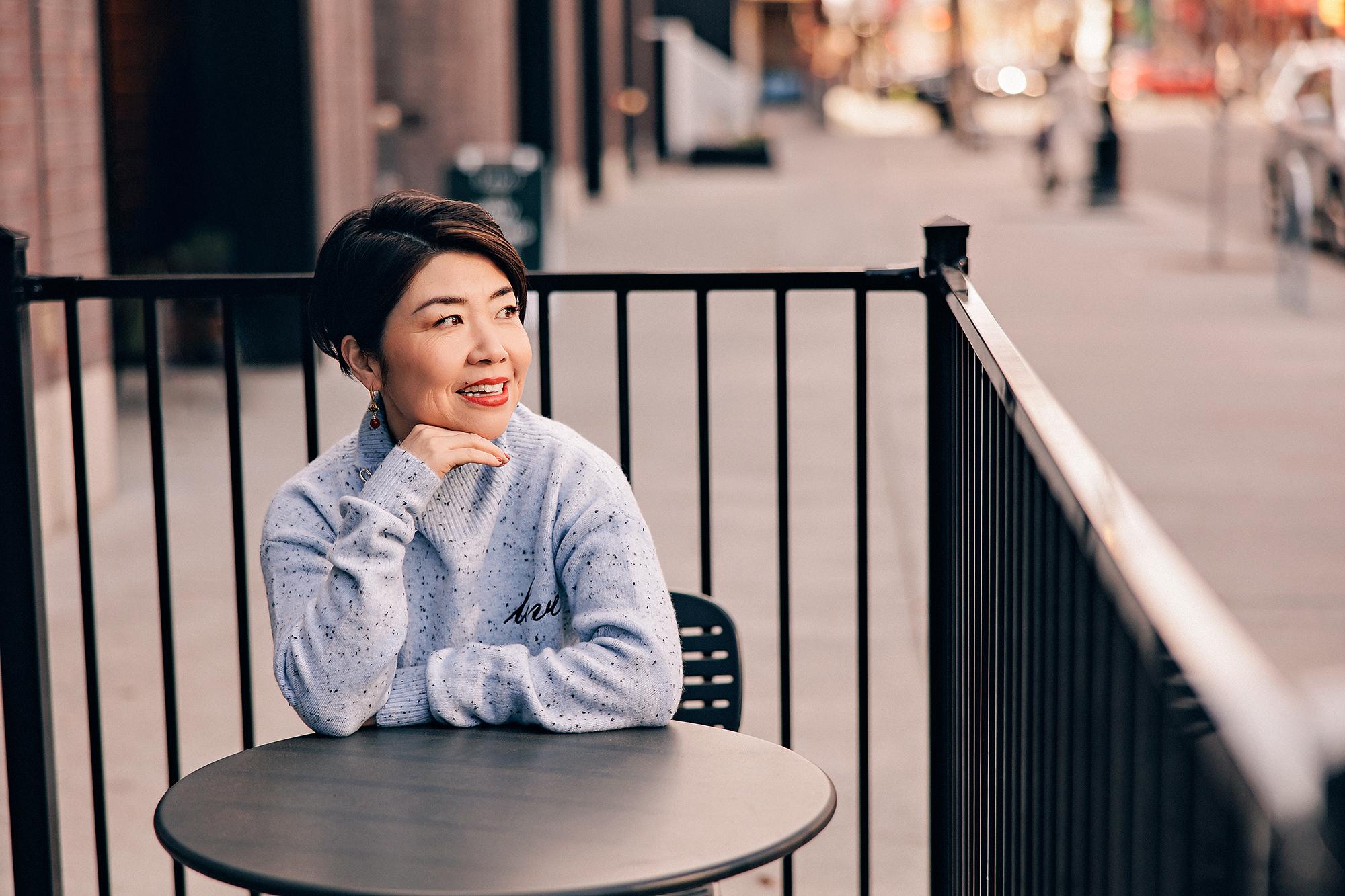 Lifestyle personal branding photography Calgary: Smiling woman with short dark hair in a grey speckled turtleneck sweater seated at a black café table on a Calgary street patio with chin resting on her hand