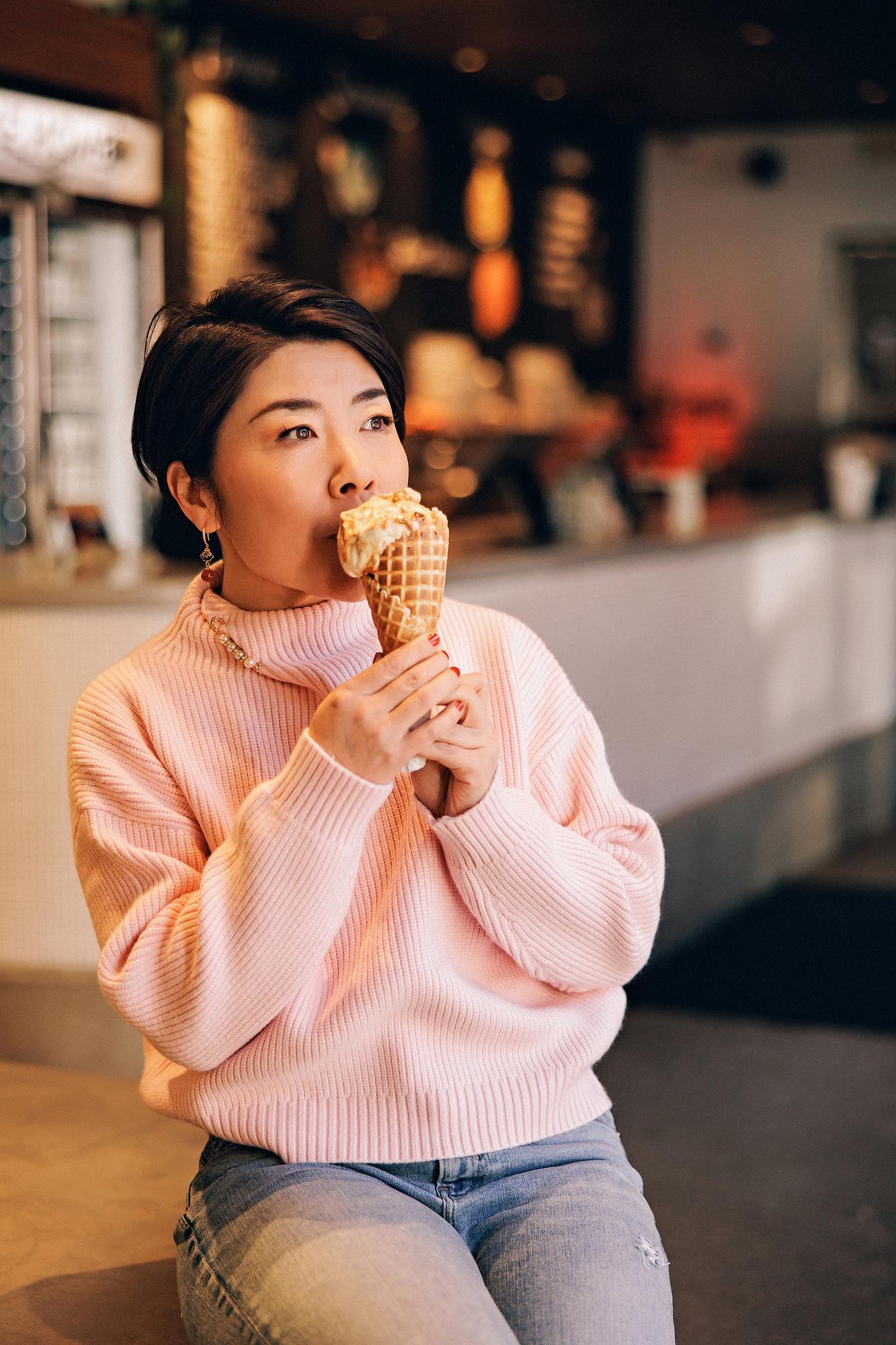 Lifestyle personal branding headshot Calgary: Candid portrait of a woman with short dark hair in a pink ribbed turtleneck sweater taking a bite of an ice cream waffle cone inside a cozy Calgary café