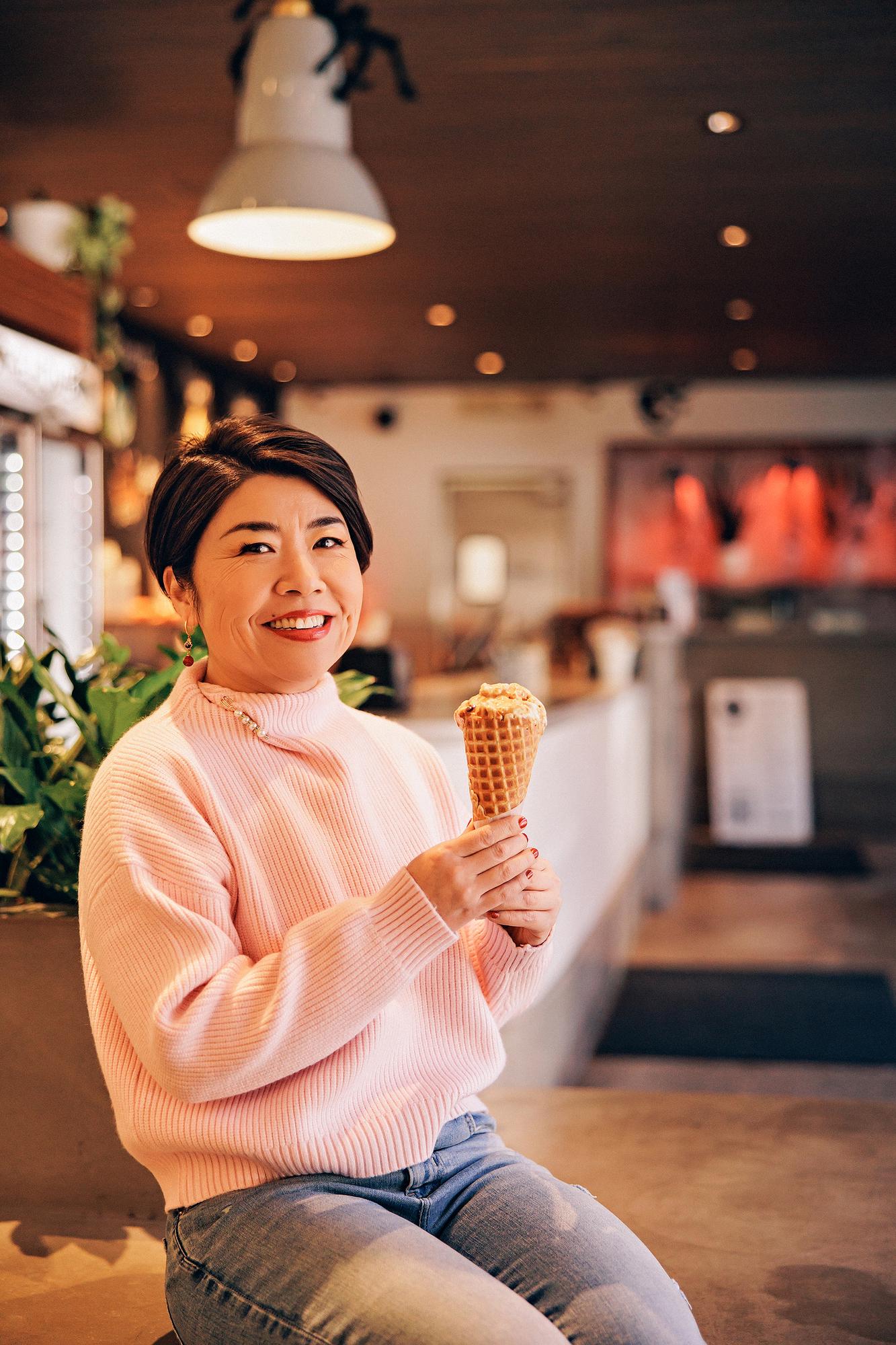 On-location personal branding photography Calgary: Smiling woman with short dark hair in a pink ribbed turtleneck holding an ice cream waffle cone while seated inside a warmly lit Calgary café with plants and pendant lighting