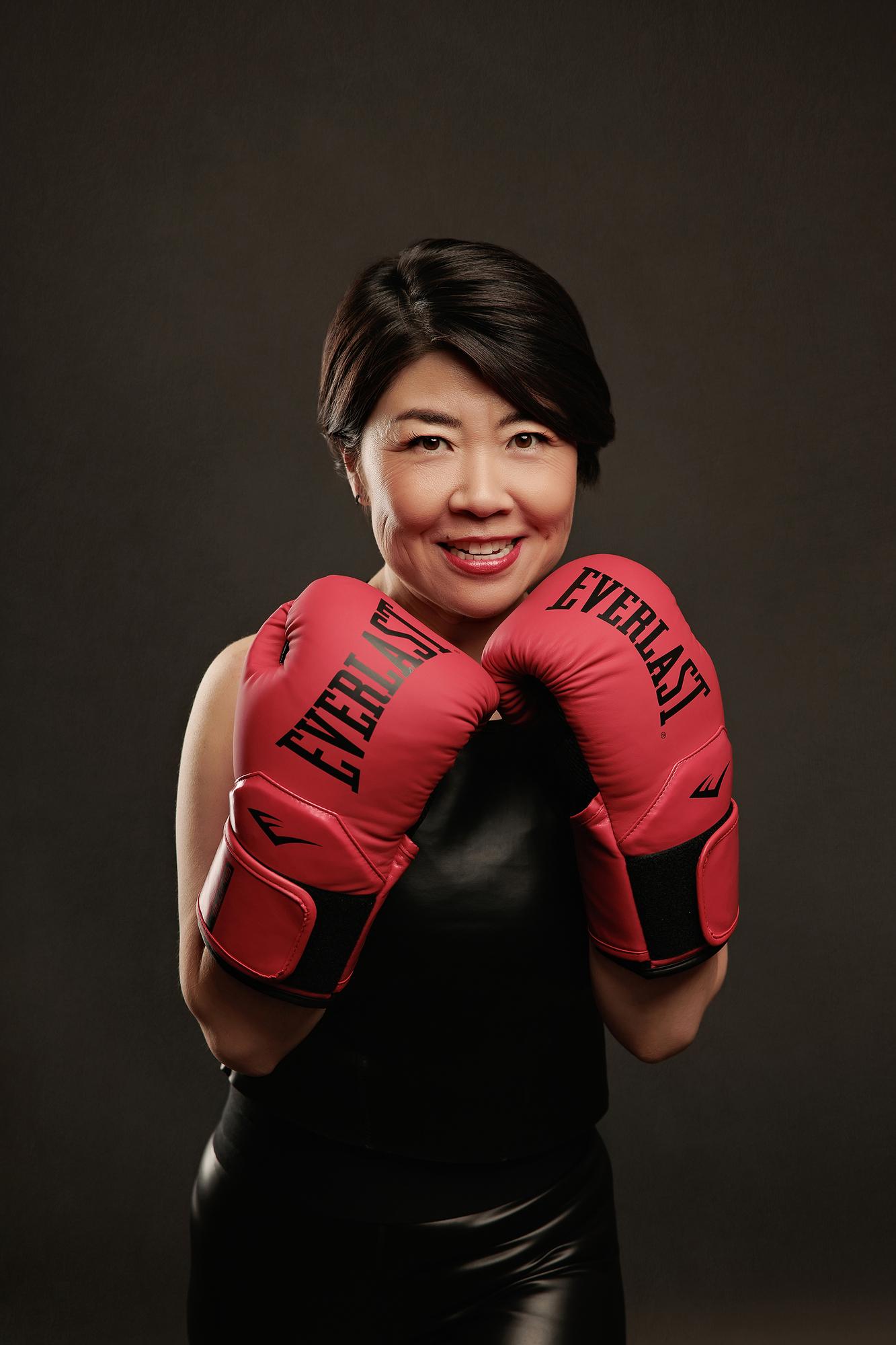 Bold personal branding headshot Calgary: Confident woman in a black sleeveless top and leather pants holding red Everlast boxing gloves in a guard position with a warm smile, dark background