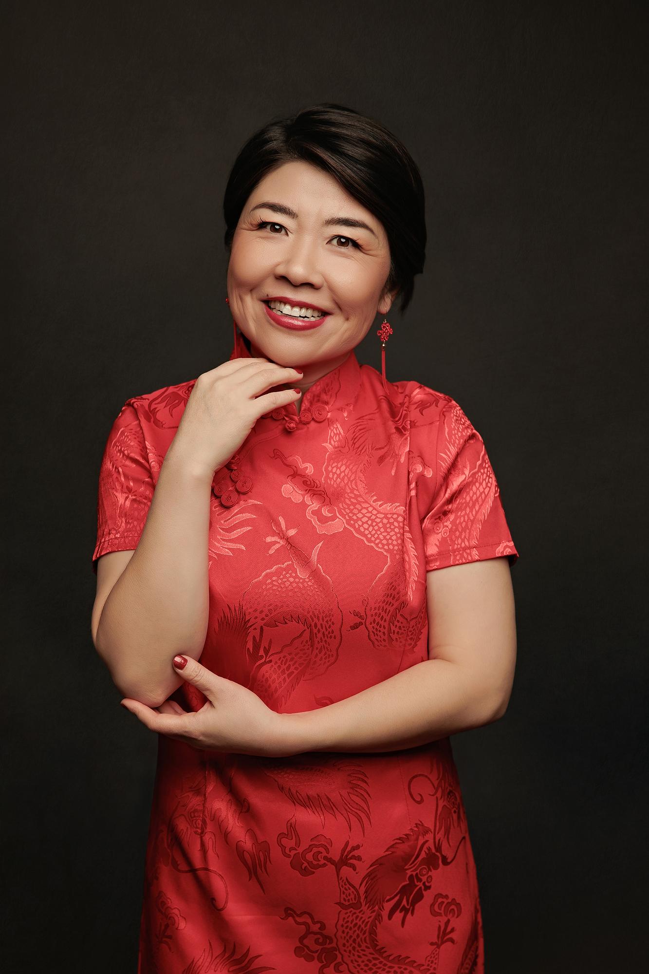 Creative personal branding photography Calgary: Radiant woman in a red dragon-print satin qipao and red tassel earrings with hand under her chin and a warm smile against a dark background