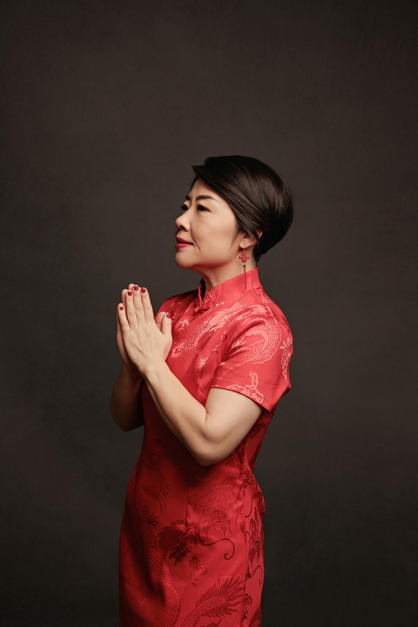 Artistic personal branding portrait Calgary: Elegant woman in a red satin qipao and red tassel earrings in a graceful side-profile pose with hands pressed together in a traditional greeting, dark background