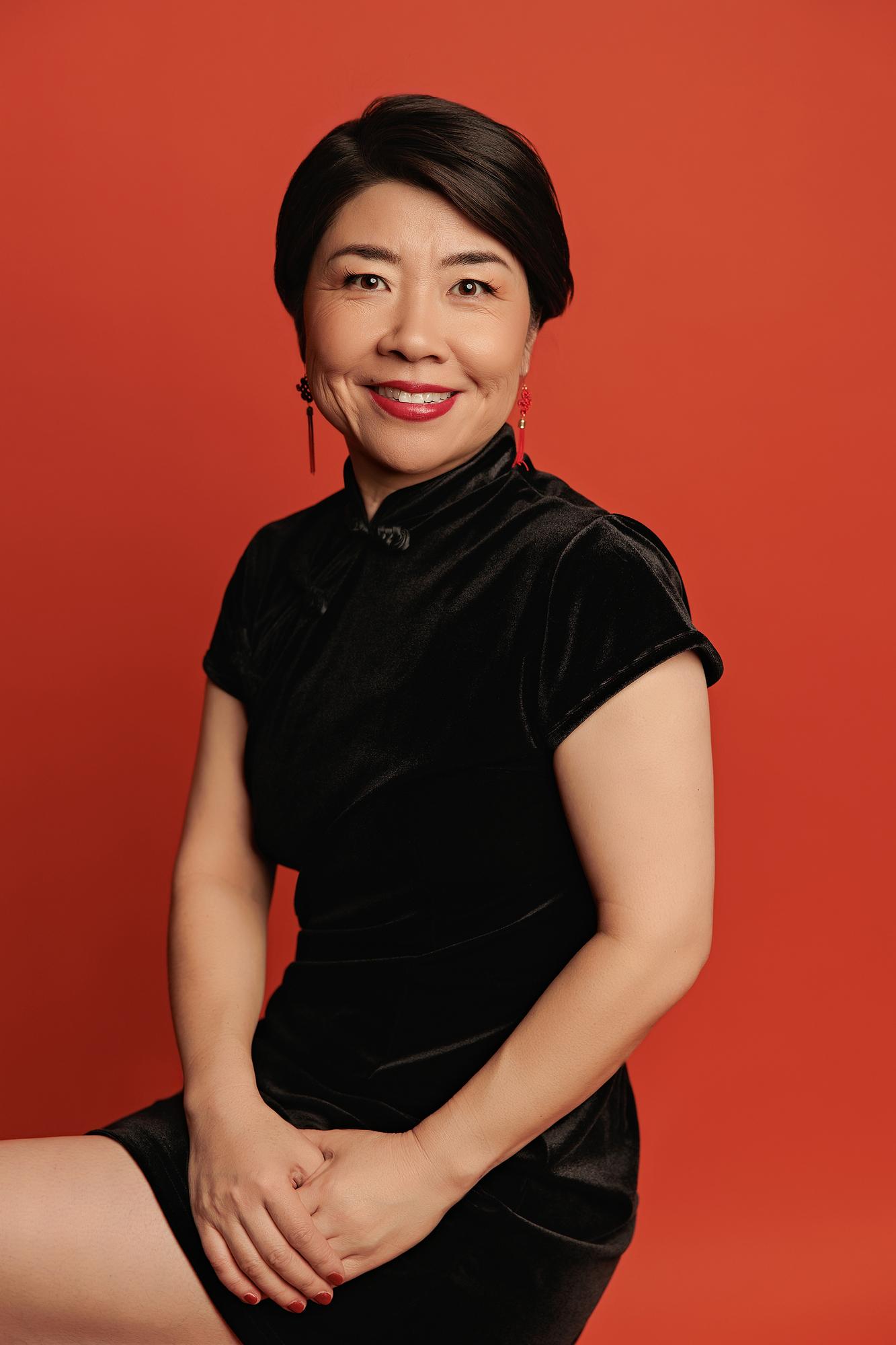 Vibrant personal branding photography Calgary: Smiling woman in a black velvet qipao and red tassel earrings seated with hands clasped against a bold red studio background