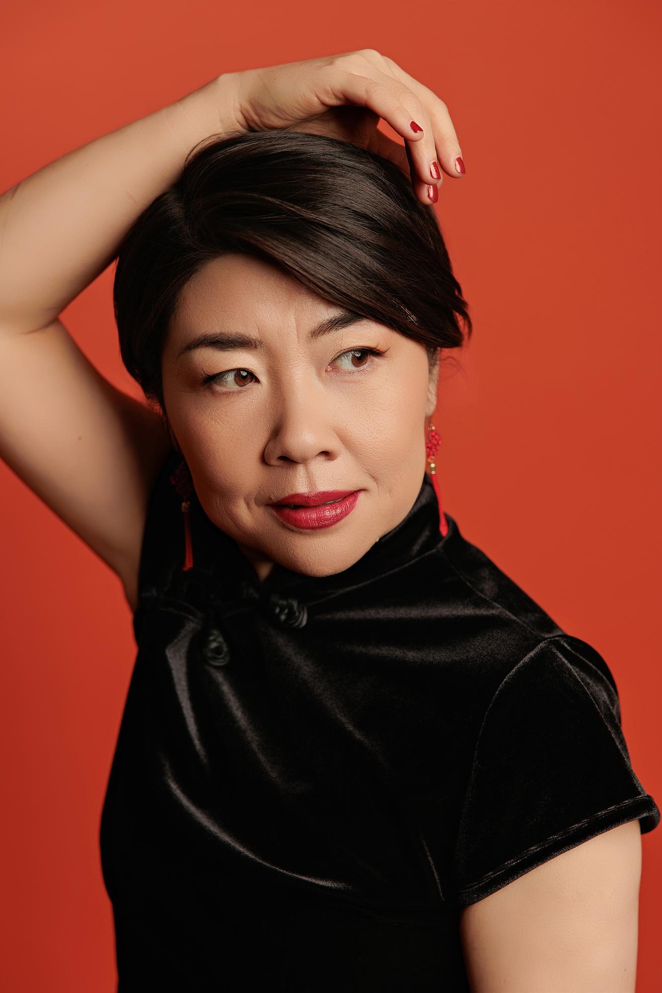 Editorial personal branding headshot Calgary: Striking close-up of a woman in a black velvet qipao and red tassel earrings with hand in hair and a direct gaze against a vivid red background