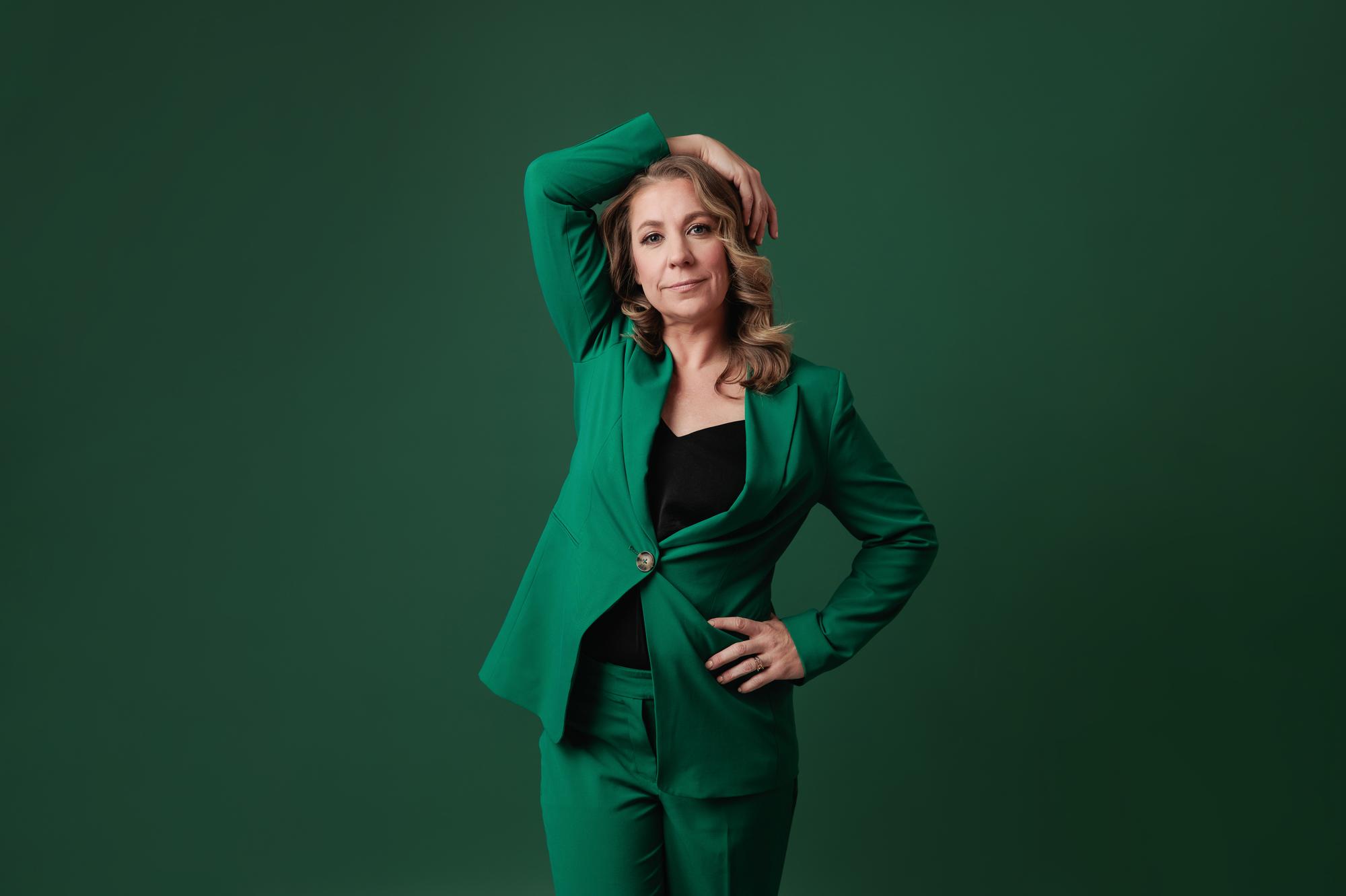 A confident woman in a vibrant emerald green pantsuit poses with her hand on her head, smiling for a professional Calgary headshot or personal branding portrait in studio.