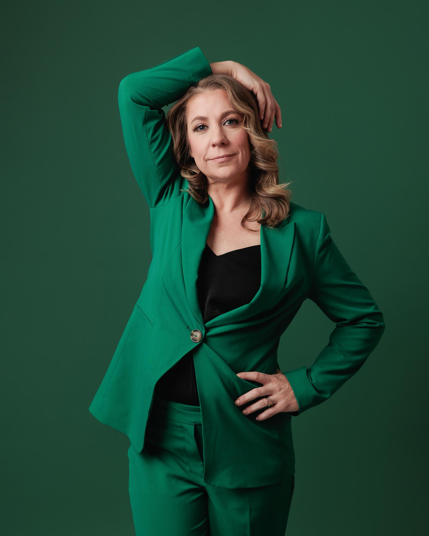 A confident woman in a vibrant green pantsuit with a subtle smile, posing against a dark green studio background, showcasing professional headshots from our Calgary photography studio.