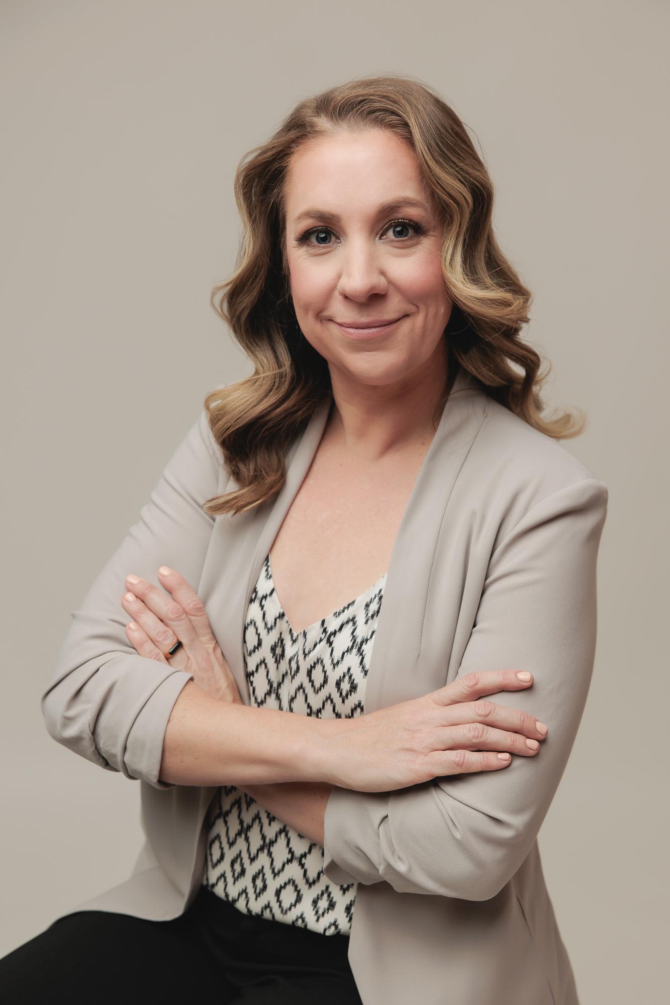 A professional woman with a friendly smile and crossed arms, wearing a grey blazer, perfectly lit for a modern headshot by a Calgary photographer.