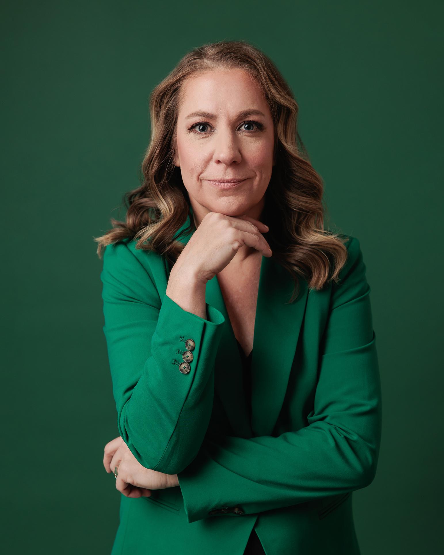 A professional woman with a confident smile, hand under chin, wears an emerald green blazer for her modern studio headshot by a Calgary photographer.