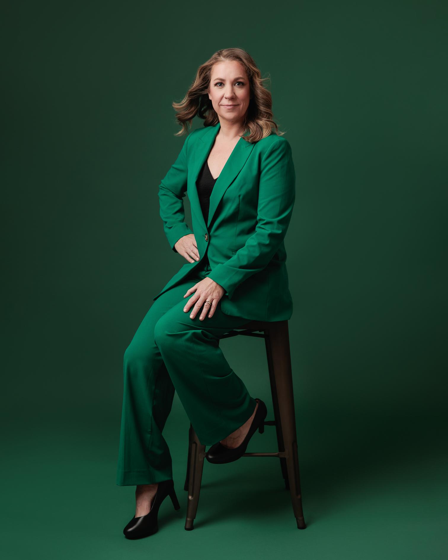 A confident woman in an emerald green pantsuit sits on a stool against a dark green background, a professional headshot created by a Calgary photographer.