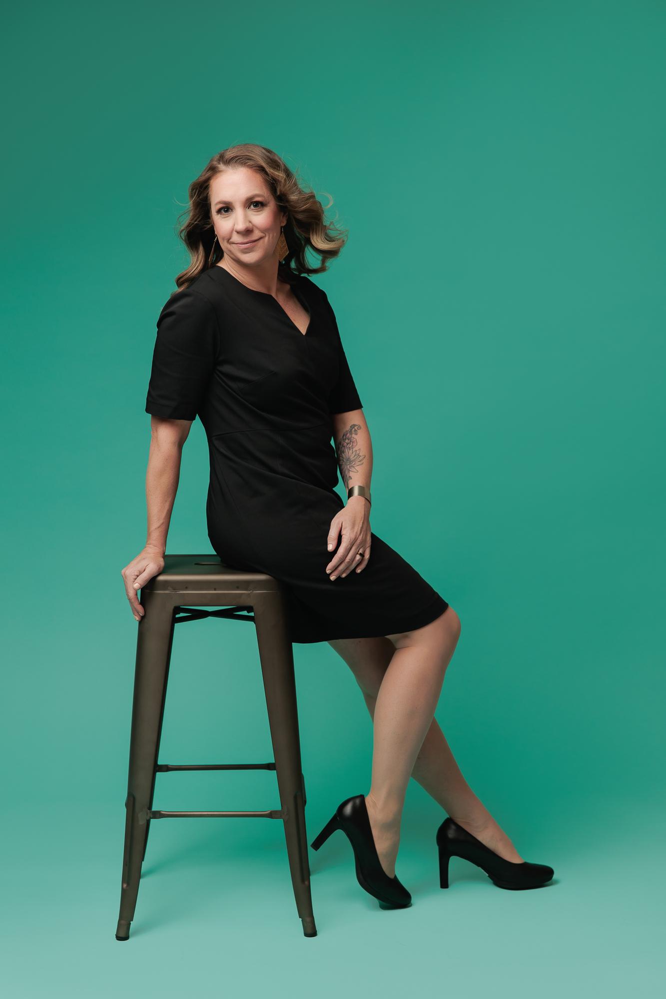 A smiling woman in a black dress with windswept hair sits on a stool against a teal backdrop, reflecting the modern professional headshots offered by our Calgary photography studio.