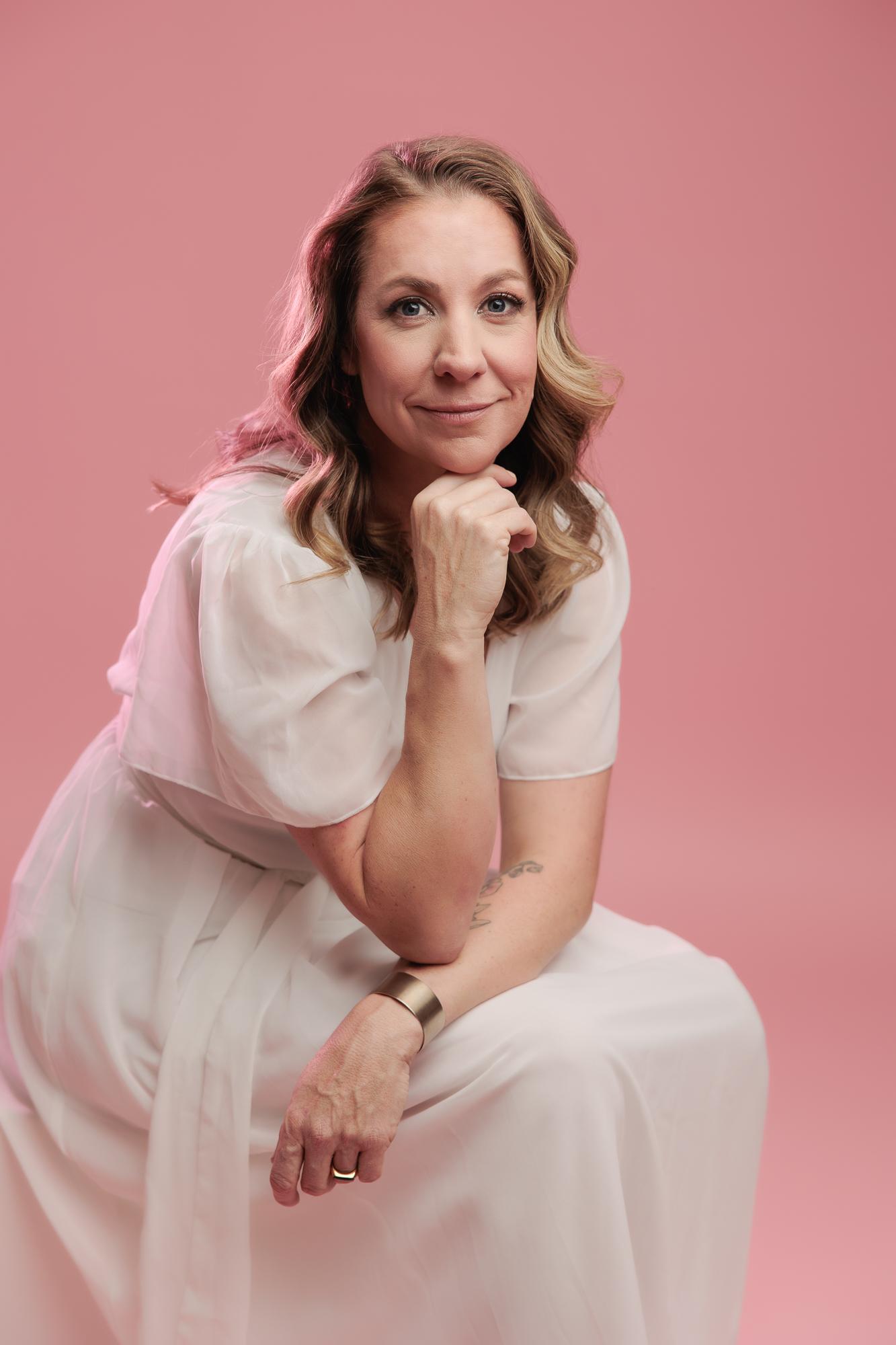 A smiling woman in a flowing white dress, with her hand gently under her chin, poses for a professional headshot at a Calgary photography studio against a vibrant pink background.