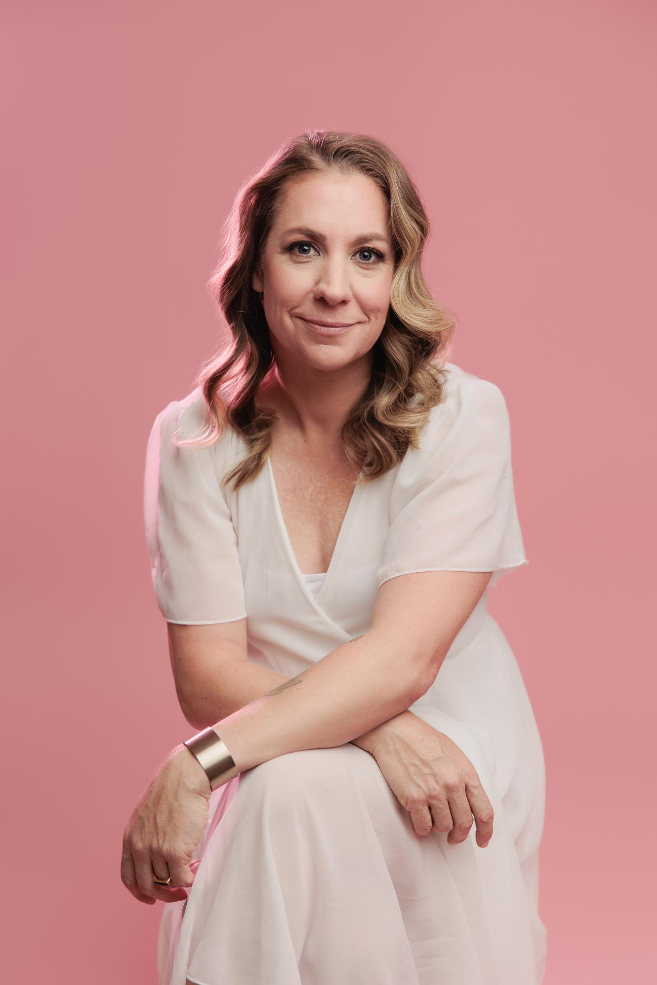 A smiling woman in a white dress poses for a personal branding session at our Calgary photography studio, featuring soft studio lighting against a pink backdrop.