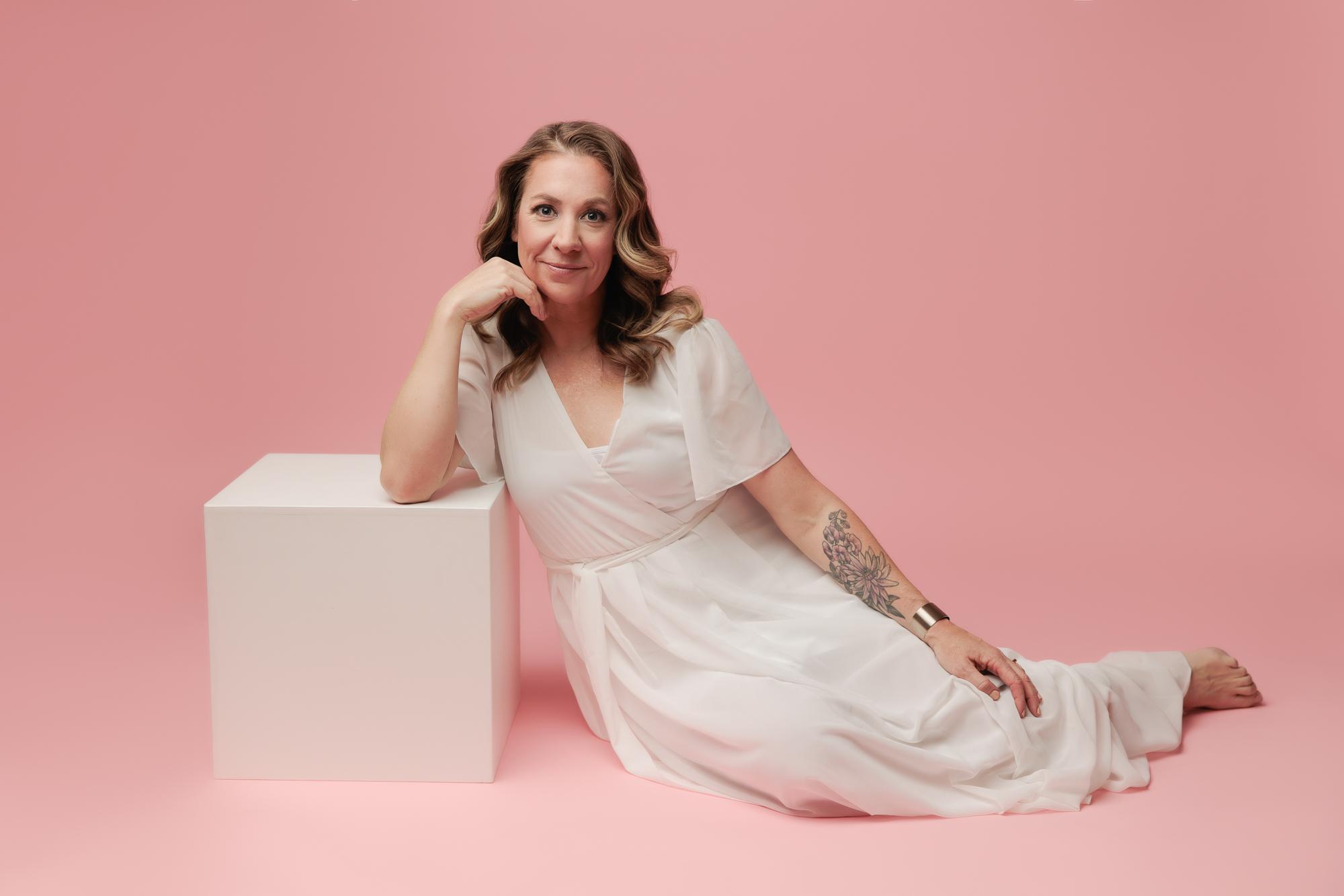 A woman in a white dress poses against a pink backdrop at our Calgary photography studio, showcasing a soft smile for her personal branding session.