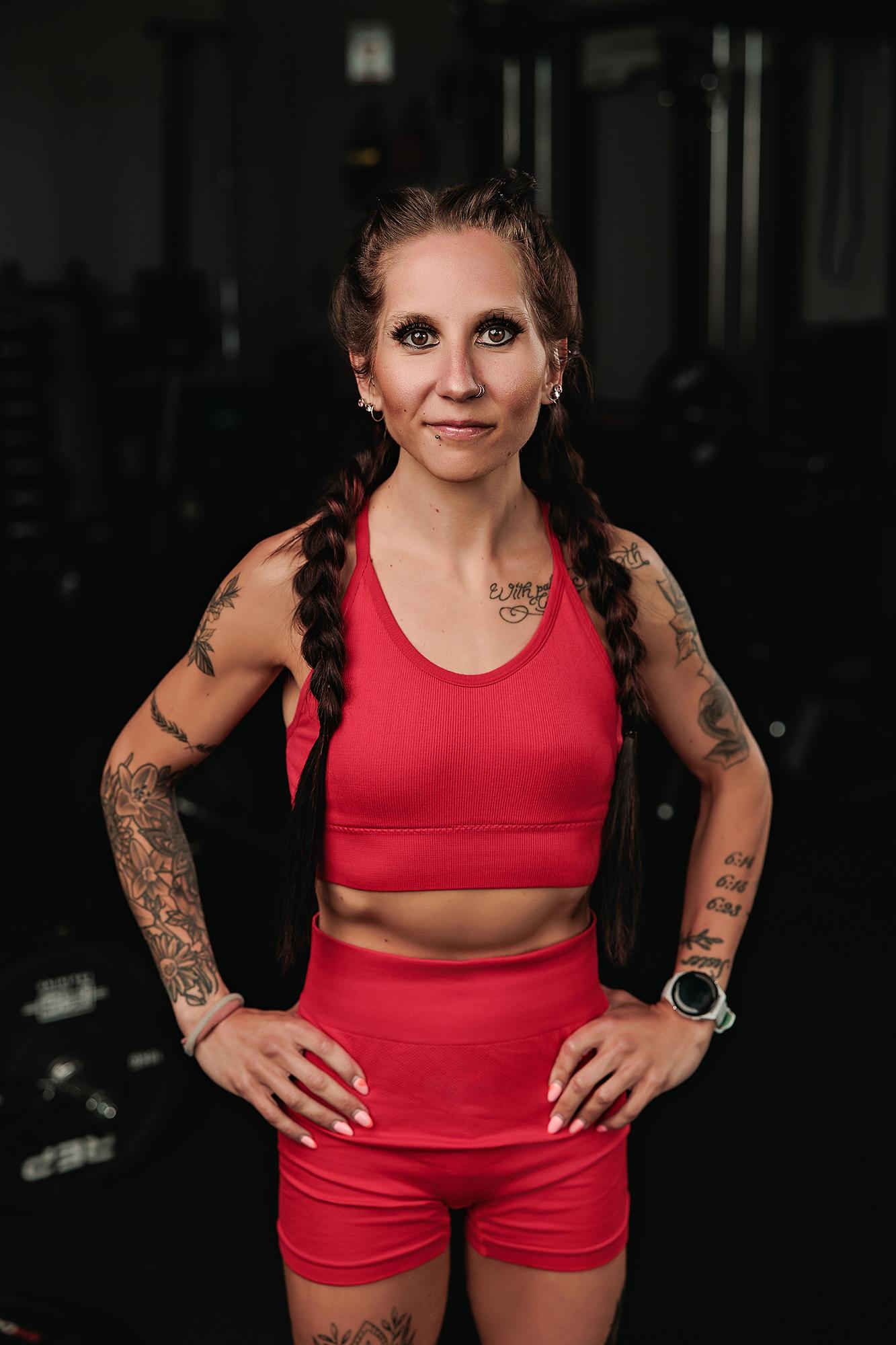 Personal branding photography Calgary gym: Confident heavily tattooed female athlete in a red ribbed sport set with hands on hips and a direct gaze, dark gym equipment behind her