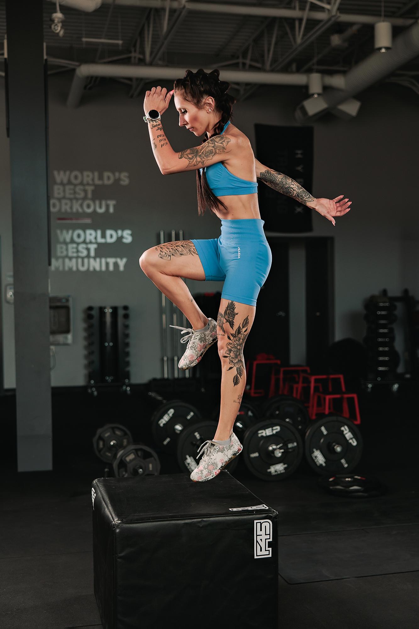 Personal branding photography Calgary gym: Tattooed female athlete in a blue sport set stepping up on a black F45 plyometric box with knee raised in an action shot, Hyrox banner and barbell plates behind her
