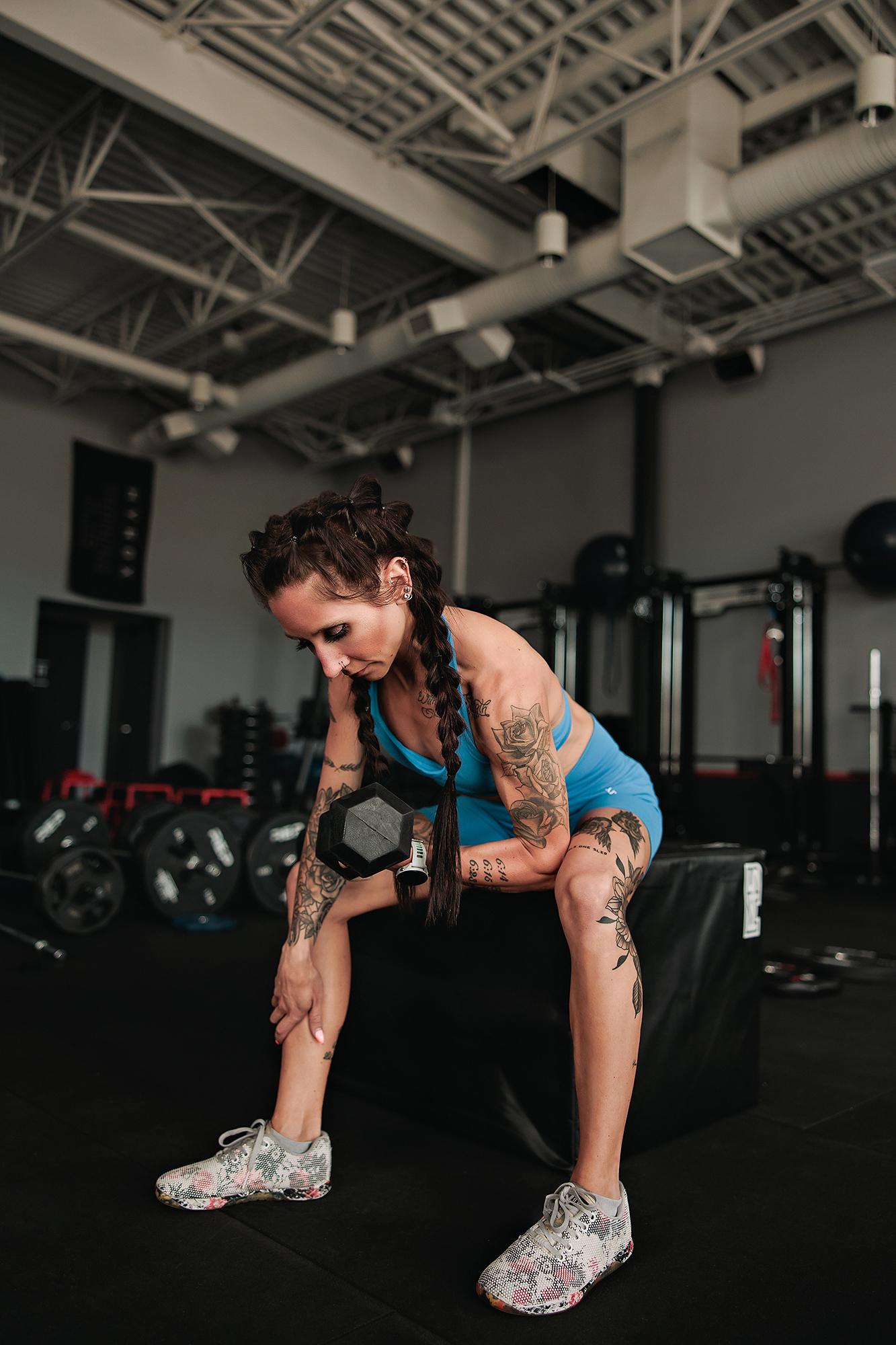 Personal branding photography Calgary gym: Full-length shot of a tattooed female athlete in a blue sport set performing a single-arm dumbbell row on a plyometric box, wide industrial gym background behind her