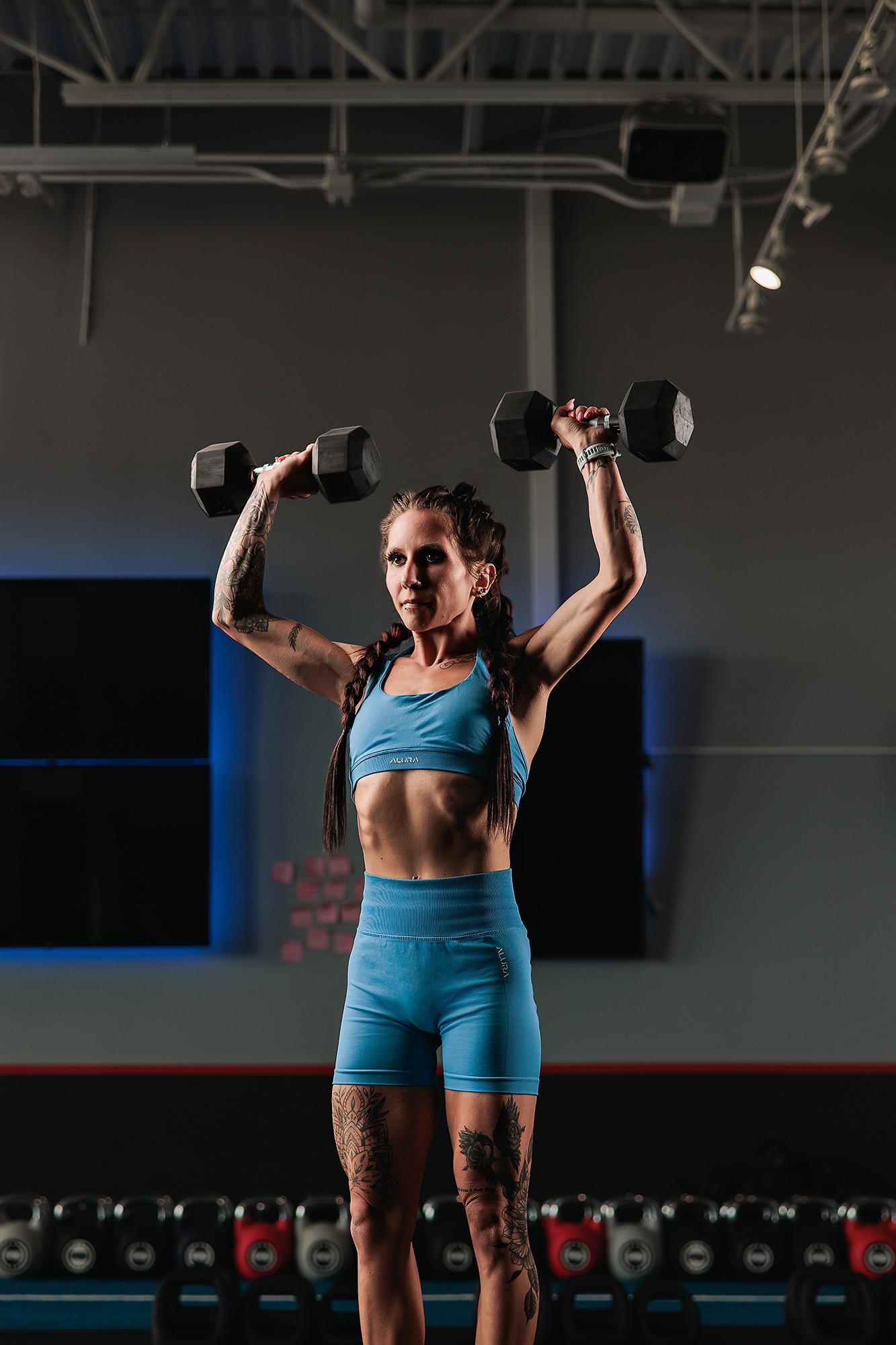 Personal branding photography Calgary gym: Tattooed female athlete in a blue sport set pressing two hex dumbbells overhead with a composed expression, wide industrial gym floor and equipment behind her