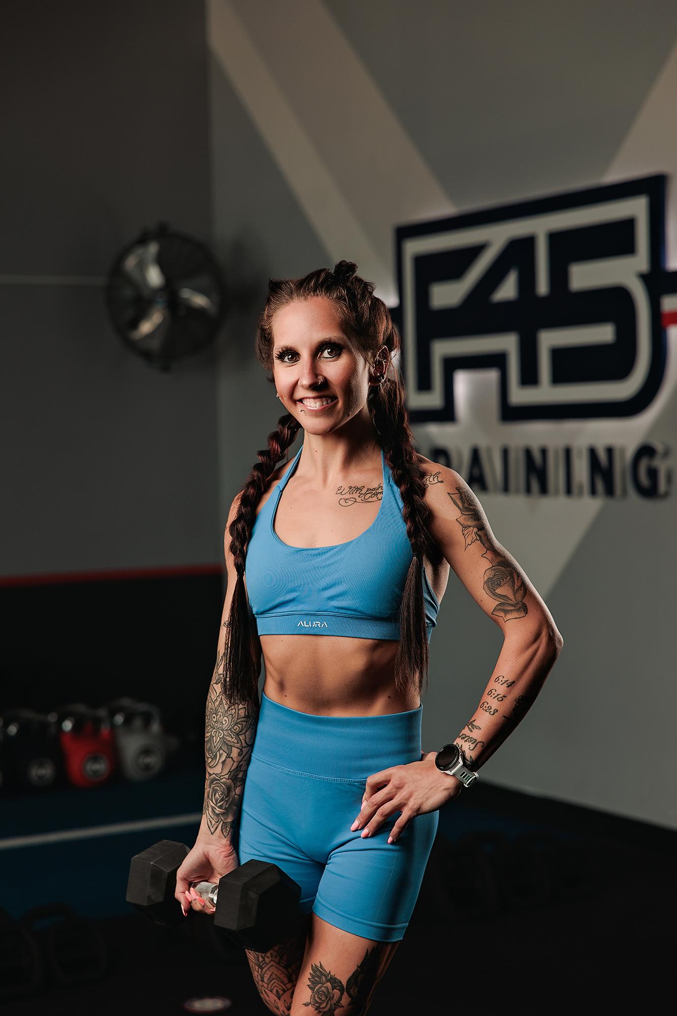 Personal branding photography Calgary gym: Tattooed female athlete in a blue ribbed sport set holding a dumbbell at her side with hand on hip and a bright smile, F45 Training logo prominently behind her