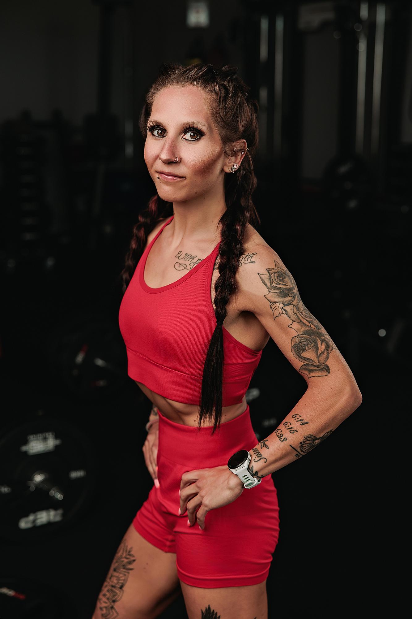 Personal branding photography Calgary gym: Tattooed female athlete in a red ribbed sport set in a three-quarter pose with hand on hip and a composed expression, dark gym background