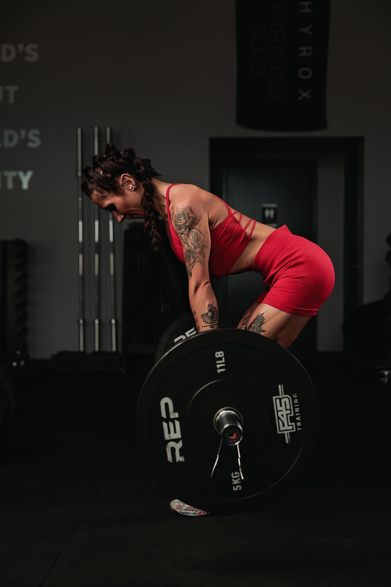 Personal branding photography Calgary gym: Tattooed female athlete in a red strappy sport bra and shorts bending over a loaded REP barbell, Hyrox Training Club banner visible in background