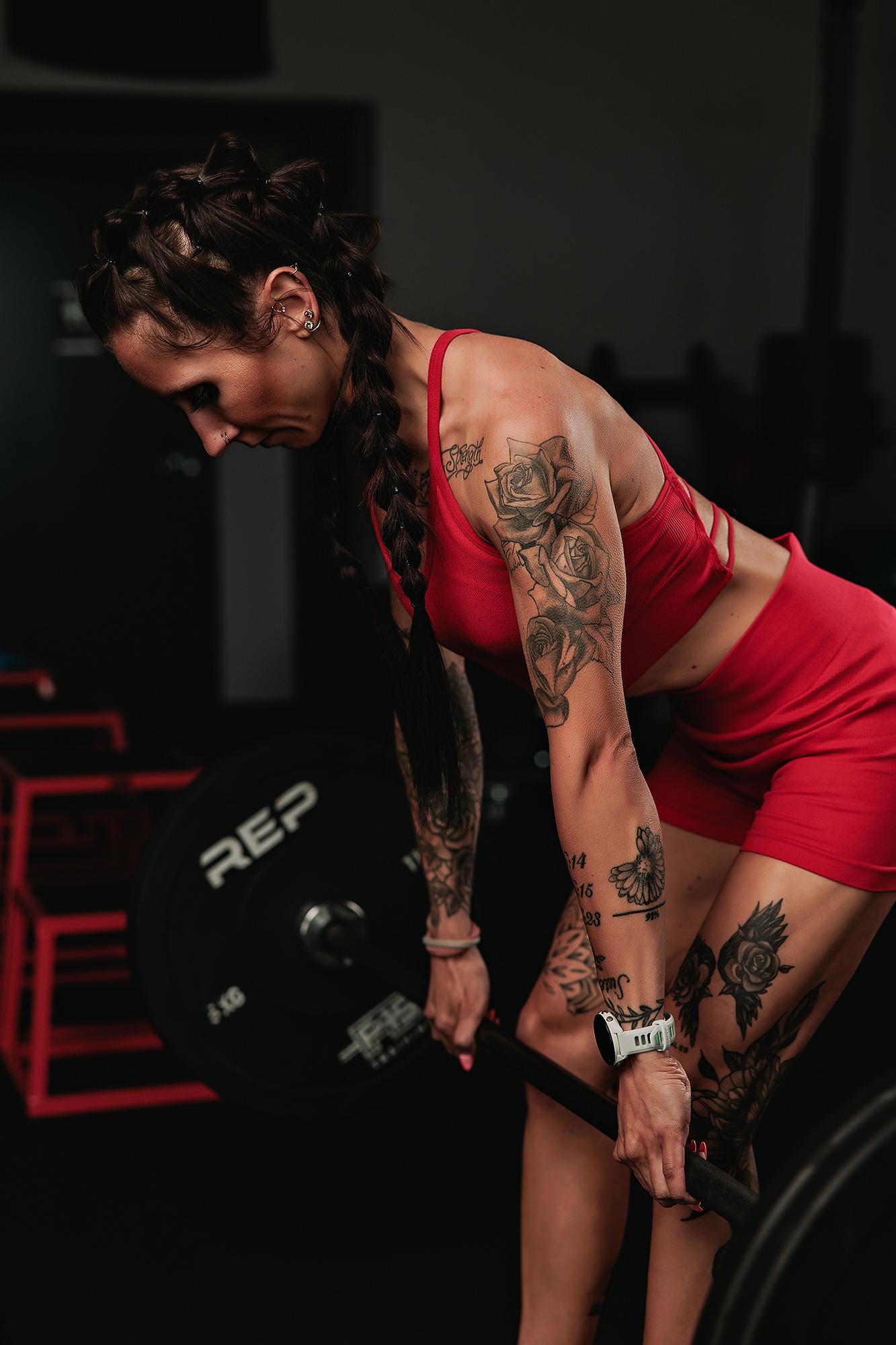 Personal branding photography Calgary gym: Close-up of a tattooed female athlete in a red strappy sport bra gripping a barbell from below, bold rose tattoos and Garmin watch visible