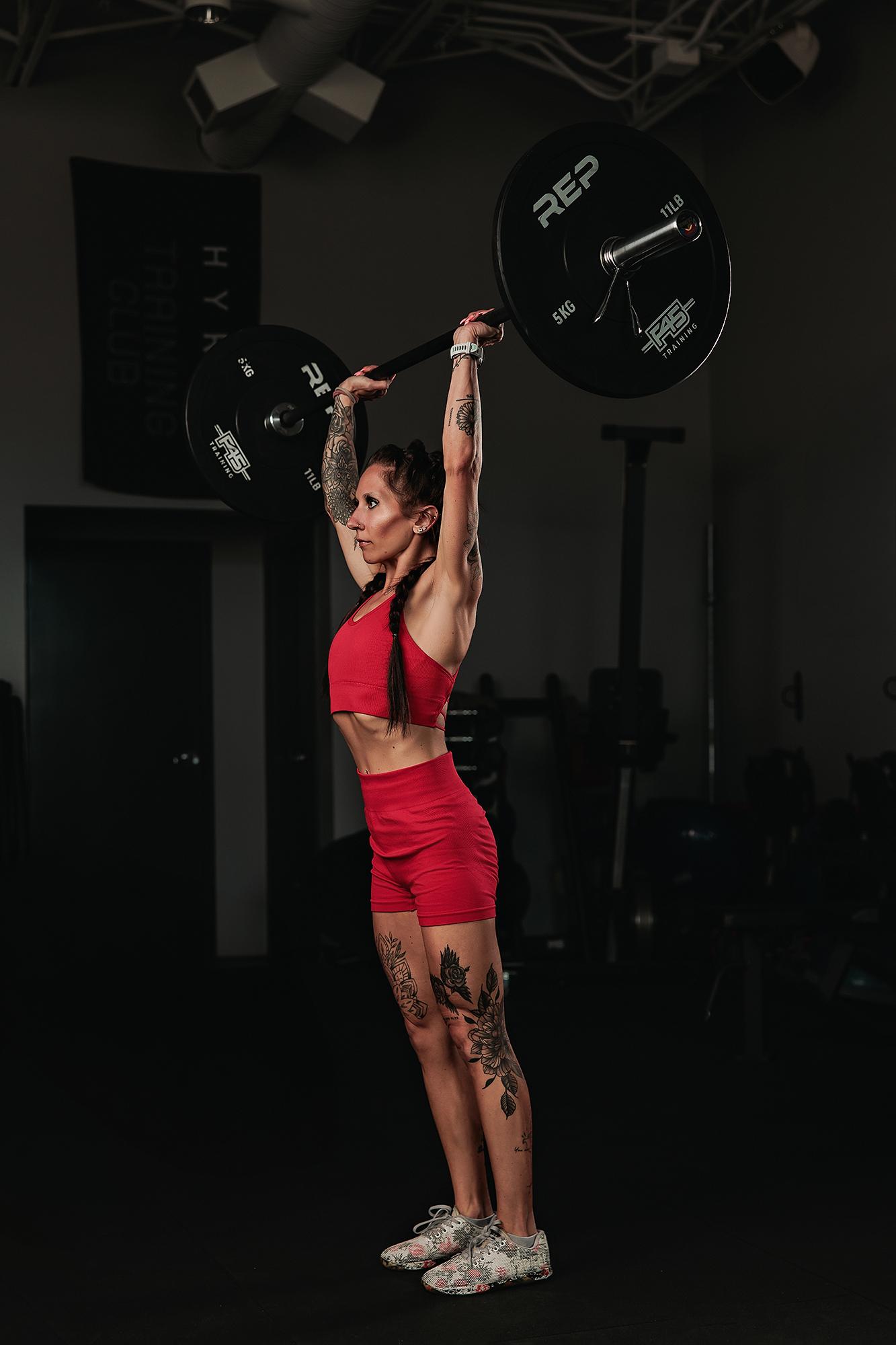 Personal branding photography Calgary gym: Tattooed female athlete in a red sport set pressing a loaded REP barbell overhead in profile, Hyrox Training Club banner and gym equipment behind her
