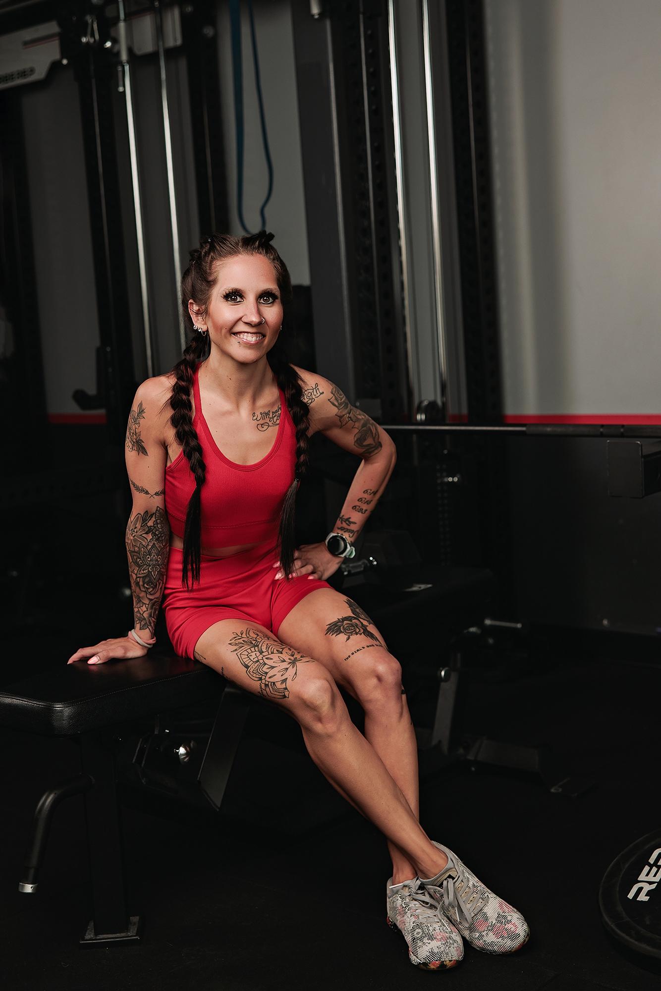 Personal branding photography Calgary gym: Tattooed female athlete in a red ribbed sport set seated on a gym bench with hand on hip and a radiant smile, cable machines behind her