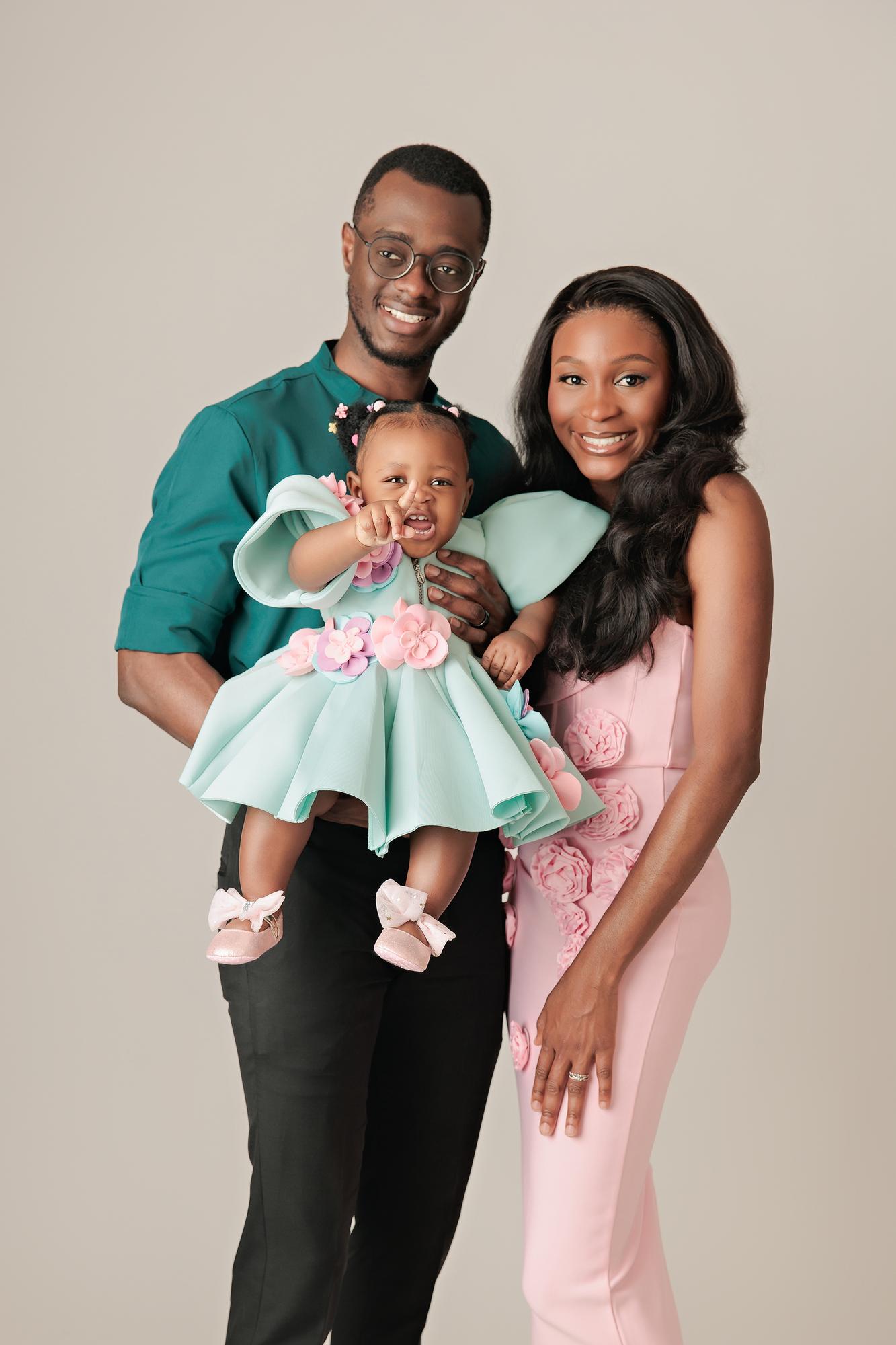 Family photography Calgary studio: Parents smiling at camera while father holds their baby girl in a mint and pink floral dress — baby reaching out playfully — mother in a blush 3D floral gown, light grey background