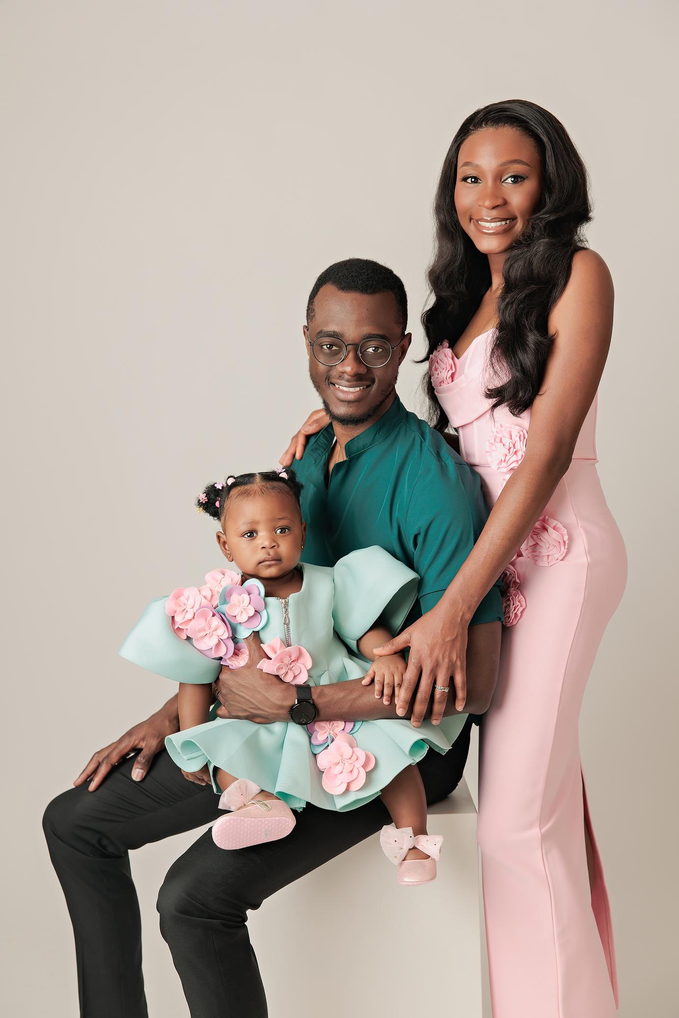 Family photography Calgary studio: Intimate portrait of parents seated together — father in a teal shirt holding their baby girl in a mint and pink floral dress, mother in a blush 3D floral gown with hand on his shoulder, light grey background
