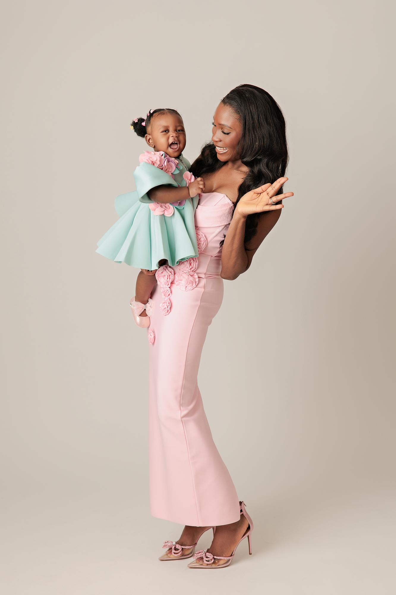 Family photography Calgary studio: Joyful mother in a blush pink 3D floral gown lifting her laughing baby girl in a mint floral dress high in the air, full-length profile view, light grey background