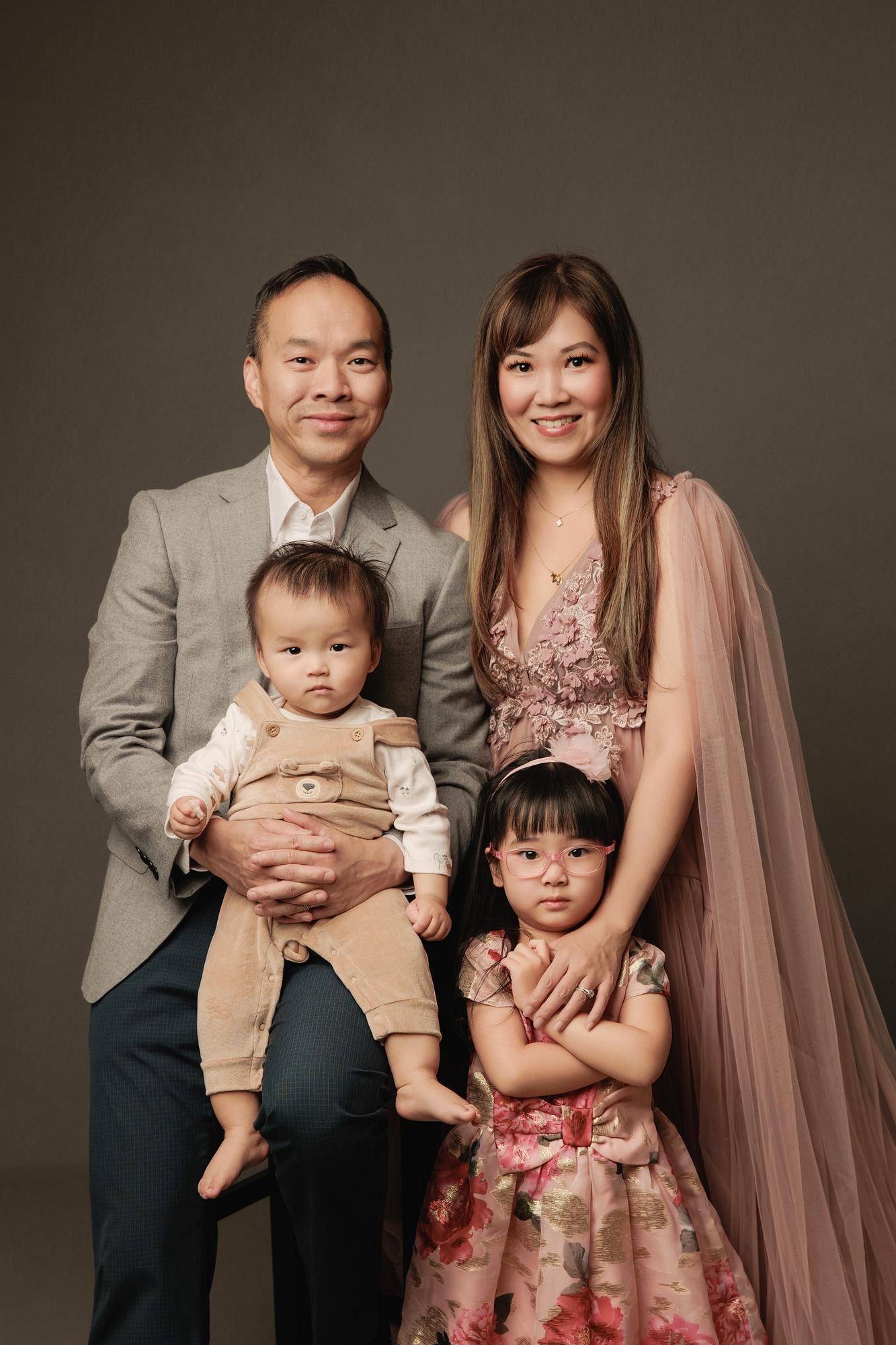 Family photography Calgary studio: Elegant family of four — father in a grey blazer holding a baby boy in a tan romper, mother in a blush pink 3D floral tulle gown, and young daughter in pink floral glasses and a brocade dress — against a warm charcoal background