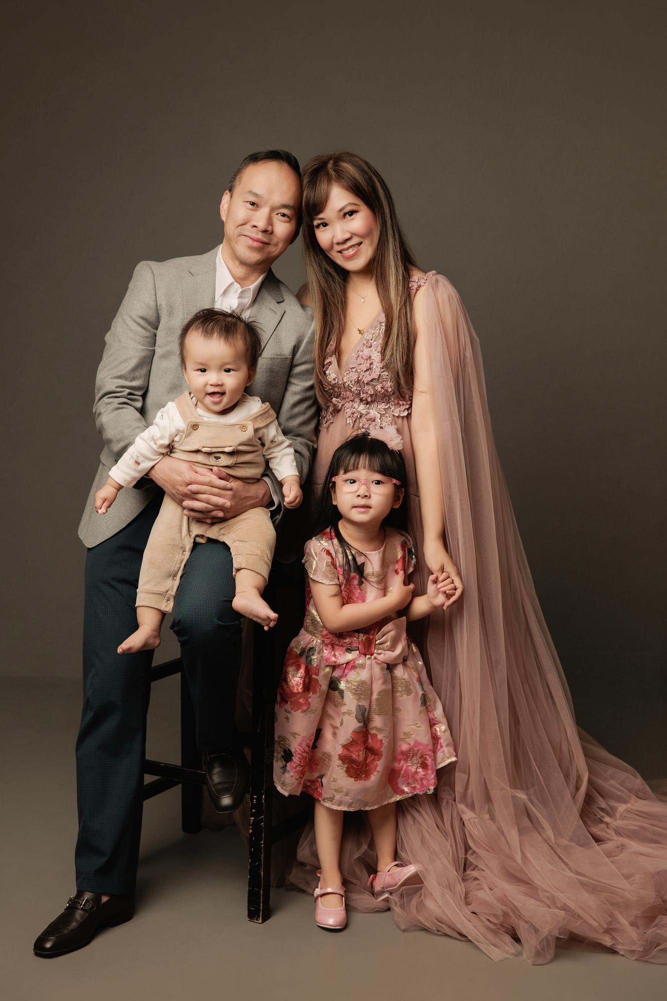 Family photography Calgary studio: Tender family portrait — father on a stool with cheek touching mother's, holding a giggling baby boy, young daughter in brocade floral dress holding mother's hand, charcoal background