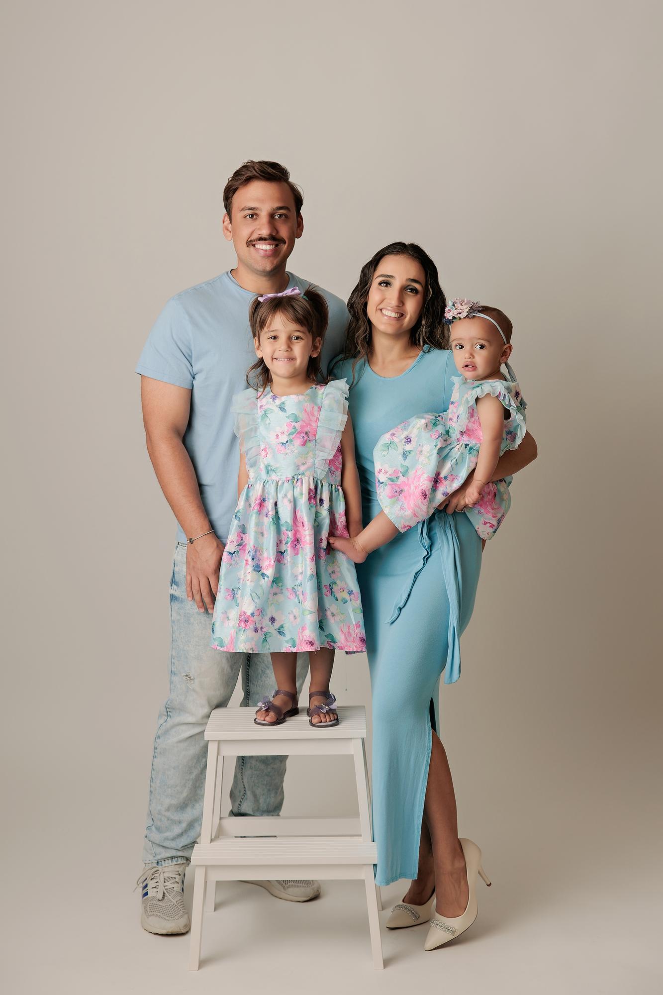 Family photography Calgary studio: Cheerful family of four in coordinated blue outfits — father in a light blue t-shirt, mother in a teal midi dress holding a baby girl in a floral dress with a flower headband, older daughter in a matching floral dress standing on a white step stool, light grey background