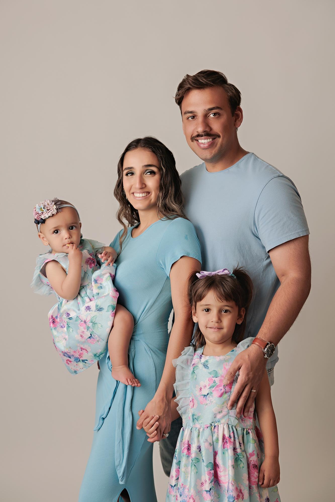 Family photography Calgary studio: Warm portrait of parents with two daughters in coordinated blue and floral outfits — mother holding baby girl with flower headband, father with arm around older daughter in floral dress, light grey background