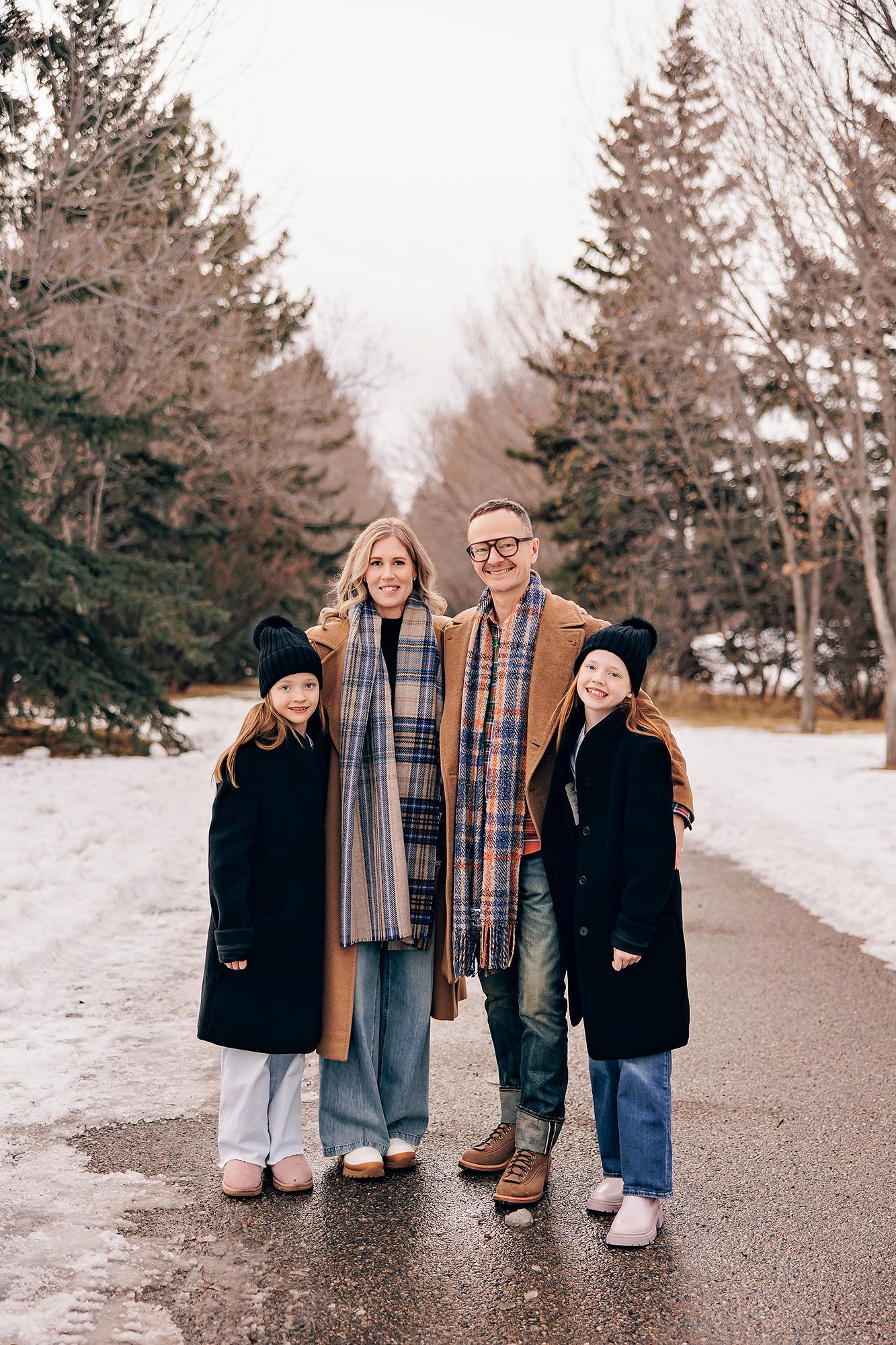 Outdoor family photography Calgary winter: Family of four in coordinated camel coats, plaid scarves, and black pom-pom toques standing on a snowy tree-lined path, surrounded by bare and evergreen trees