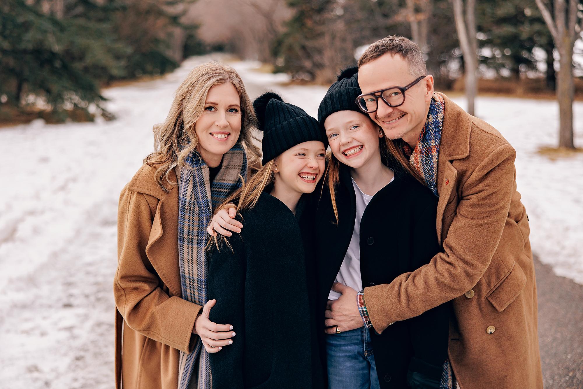 Outdoor family photography Calgary winter: Laughing family of four in camel coats and black toques — parents hugging two daughters close on a snowy Calgary park path, bare trees in background