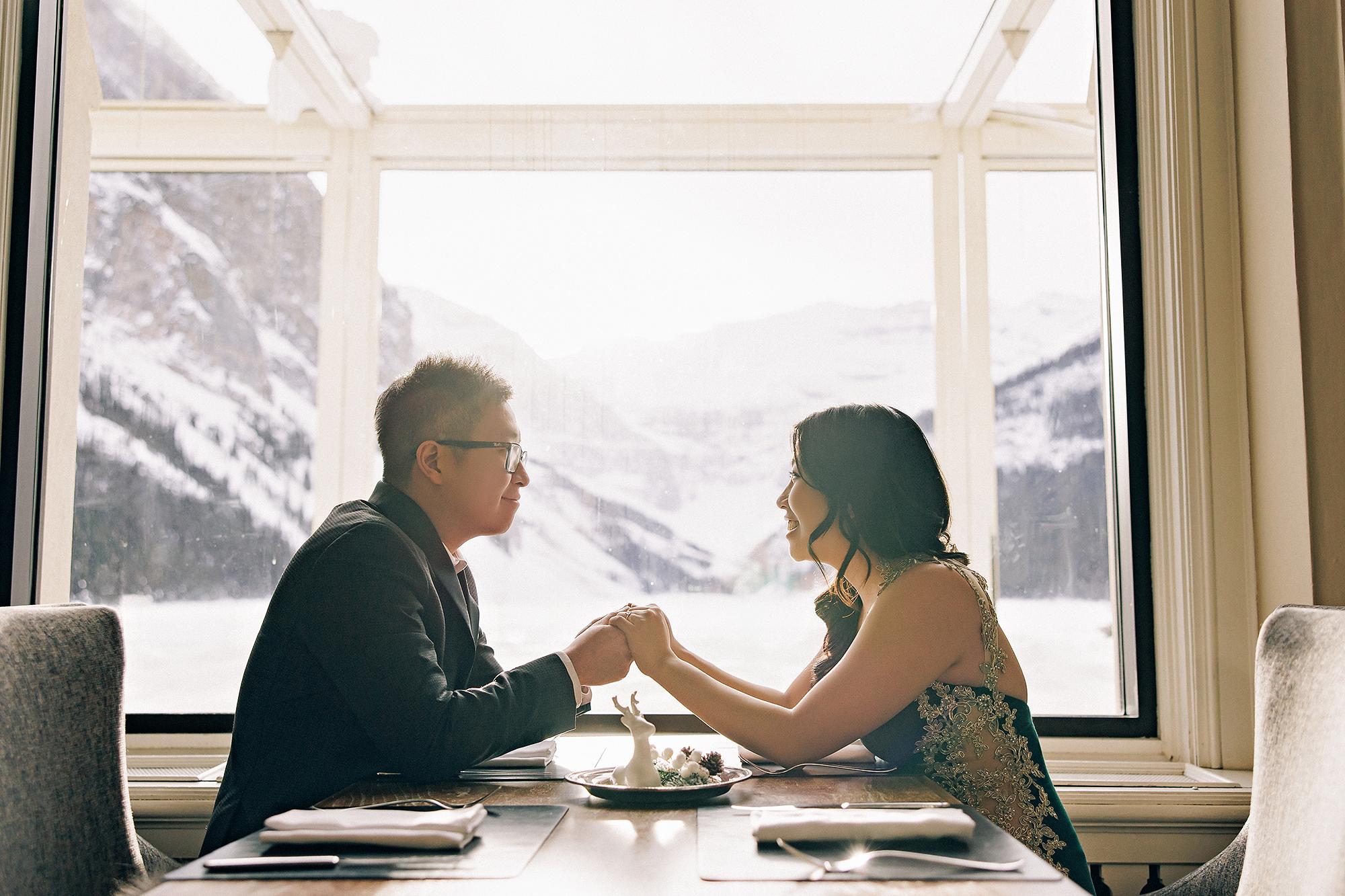 Fairmont Lake Louise engagement photography: Couple holding hands across a fine dining table — man in dark suit and woman in an emerald gold-lace gown — with dramatic snow-covered mountains and frozen lake visible through floor-to-ceiling windows behind them