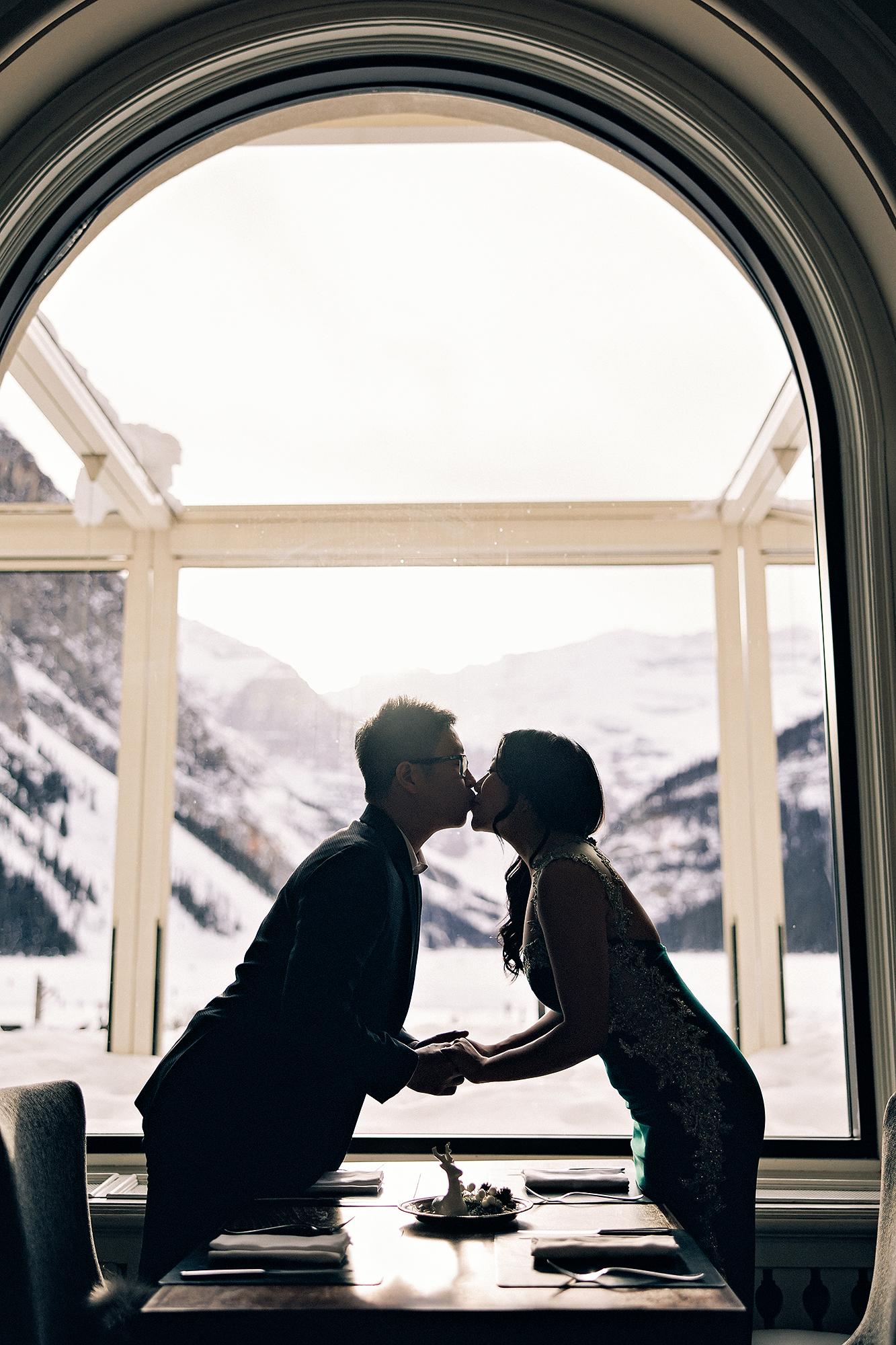 Fairmont Lake Louise engagement photography: Romantic silhouette of couple kissing across a fine dining table, framed by a grand arched window with snow-capped mountains and frozen lake glowing behind them