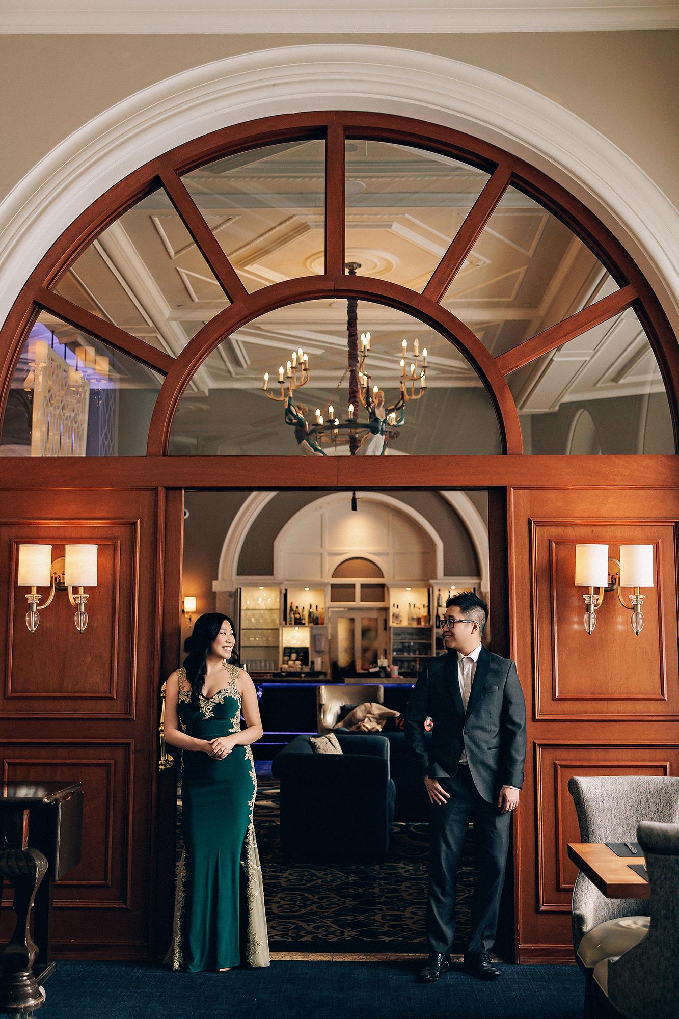 Fairmont Lake Louise engagement photography: Elegant couple standing on either side of a grand arched wood-and-glass doorway in the hotel bar — woman in an emerald and gold-lace gown, man in a dark suit — chandelier and sconce lighting above them