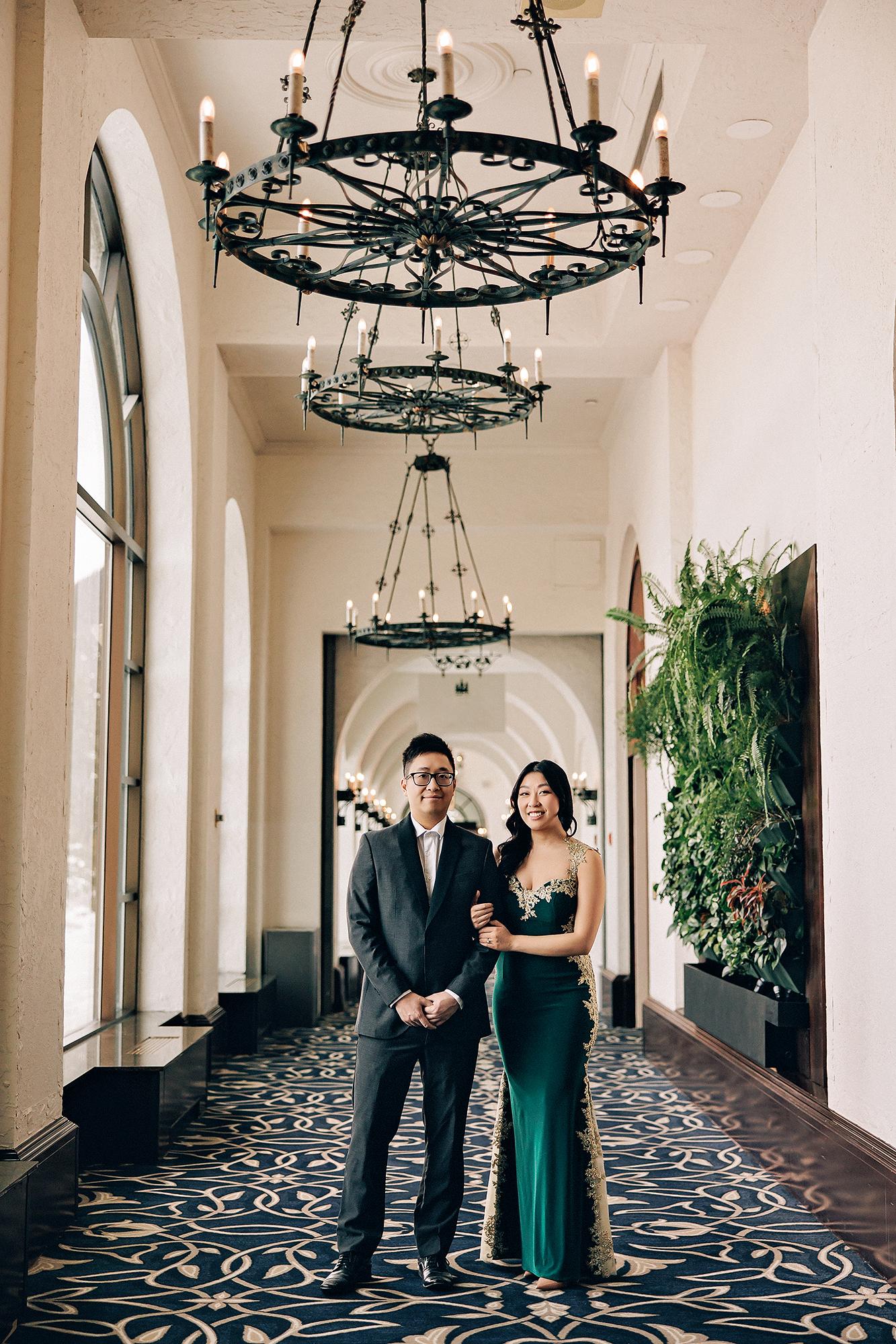 Fairmont Lake Louise engagement photography: Couple standing arm-in-arm in a grand hotel corridor lined with arched windows and ornate iron chandeliers — man in dark suit, woman in emerald and gold-lace gown — living plant wall beside them