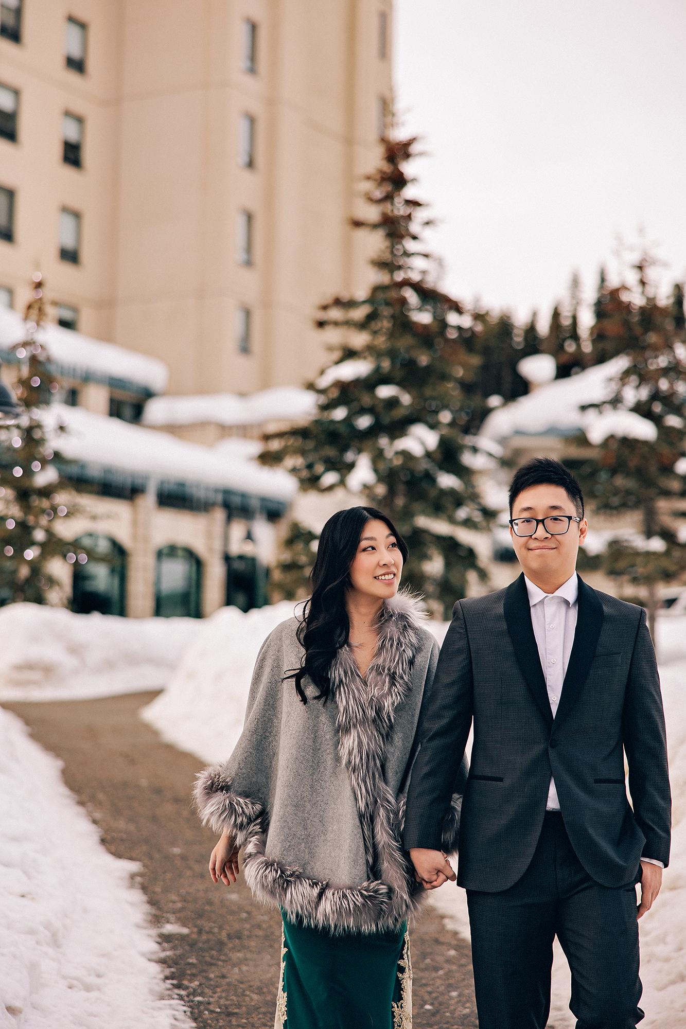 Fairmont Lake Louise engagement photography: Couple walking hand-in-hand along a snowy path outside the Fairmont Lake Louise — woman in an emerald gold-lace gown with a grey fur-trim cape, man in a dark charcoal suit — snow-laden evergreens and the hotel tower behind them