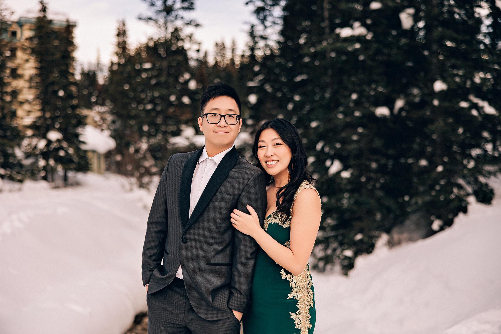 Lake Louise winter engagement photography: Couple embracing and smiling at camera in a snowy landscape — woman in an emerald and gold-lace gown, man in a dark charcoal suit — snow-covered evergreens and the Fairmont tower softly bokeh behind them