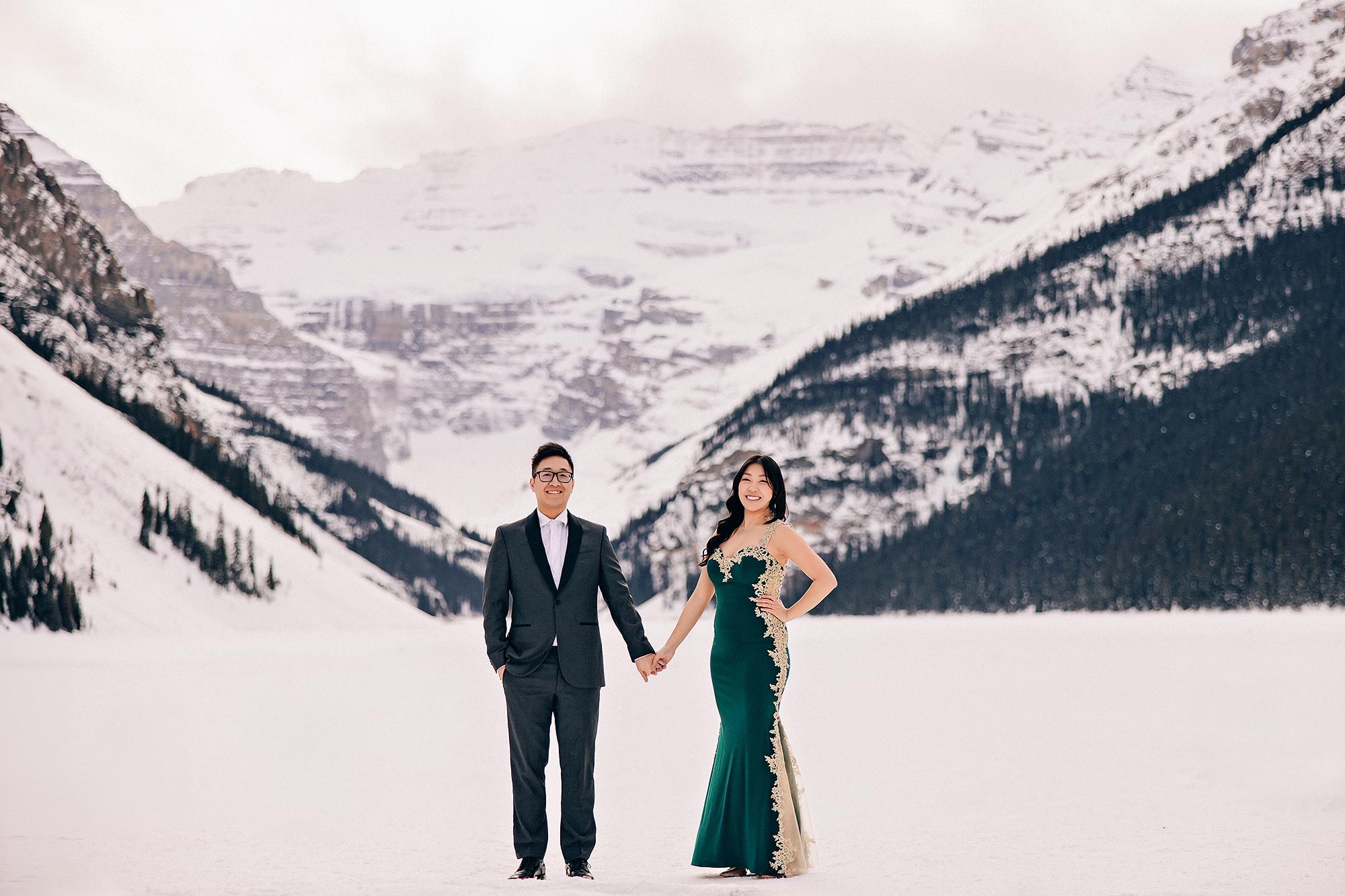 Lake Louise winter engagement photography: Full-length portrait of couple standing hand-in-hand on the frozen lake — man in dark charcoal tuxedo suit, woman in emerald and gold-lace mermaid gown — dramatic snow-covered mountain valley stretching behind them